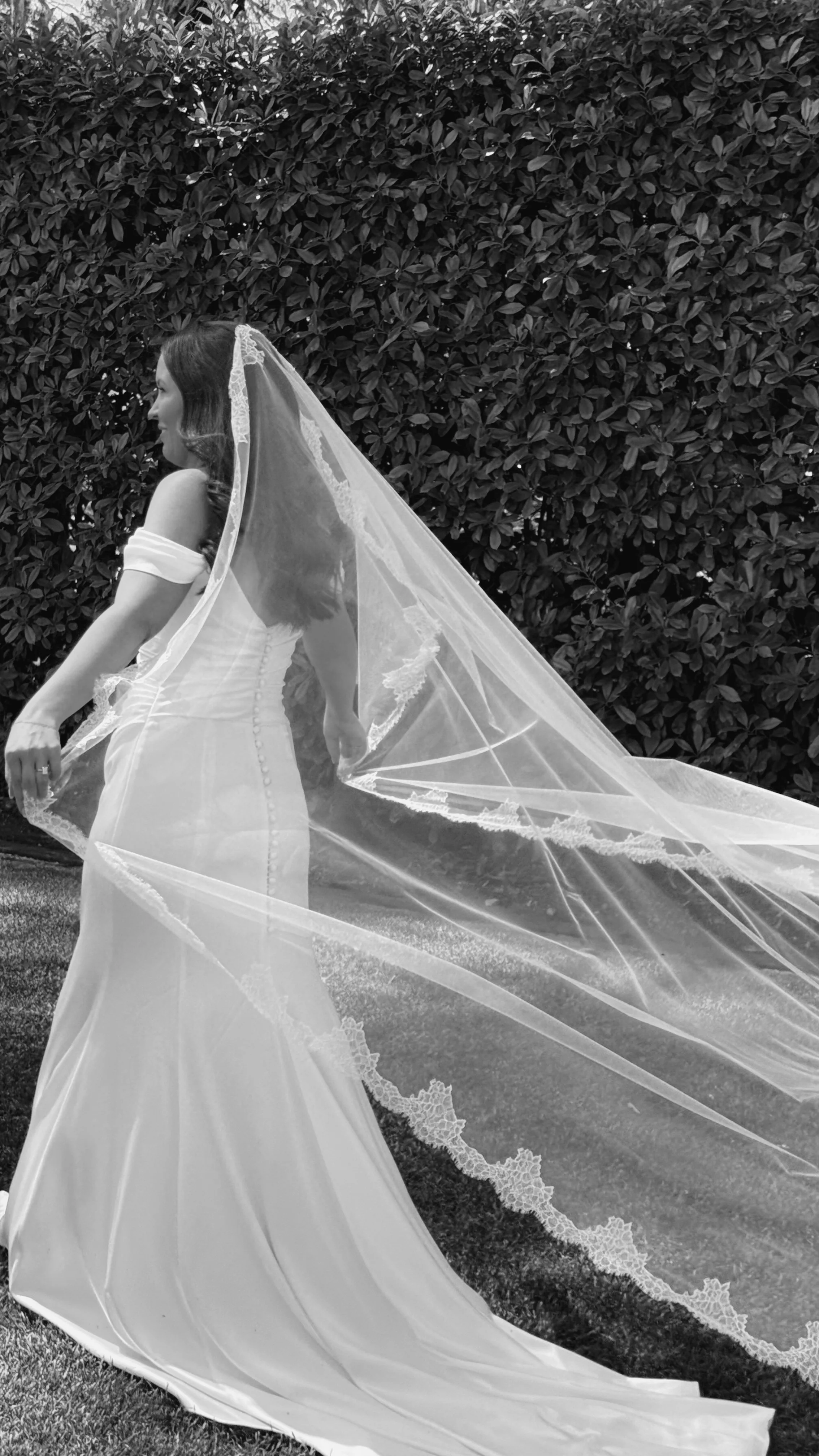 A bride in a wedding dress and veil standing outdoors in front of a leafy bush, facing left.