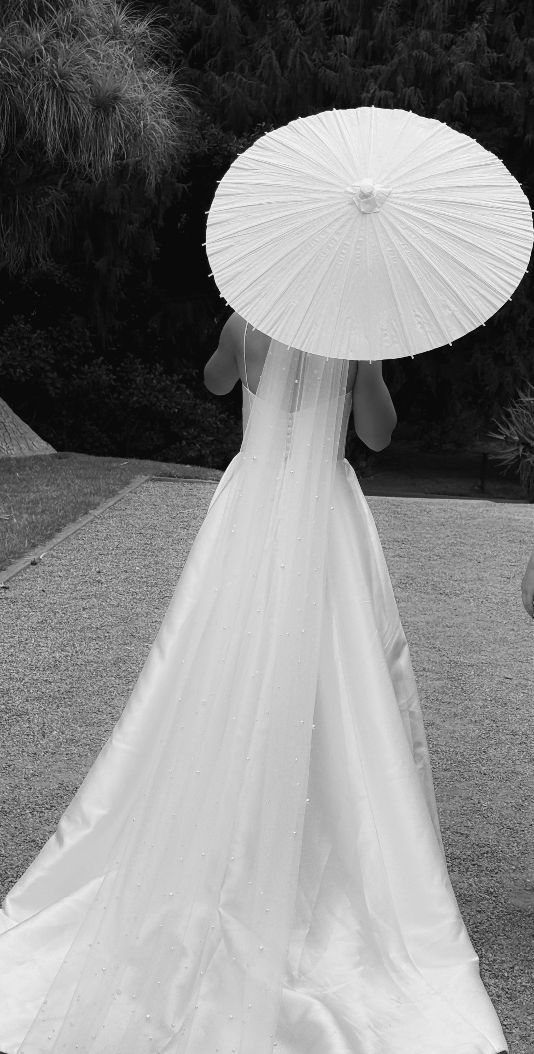 A woman in a wedding dress walking outdoors, holding a parasol, with trees in the background.