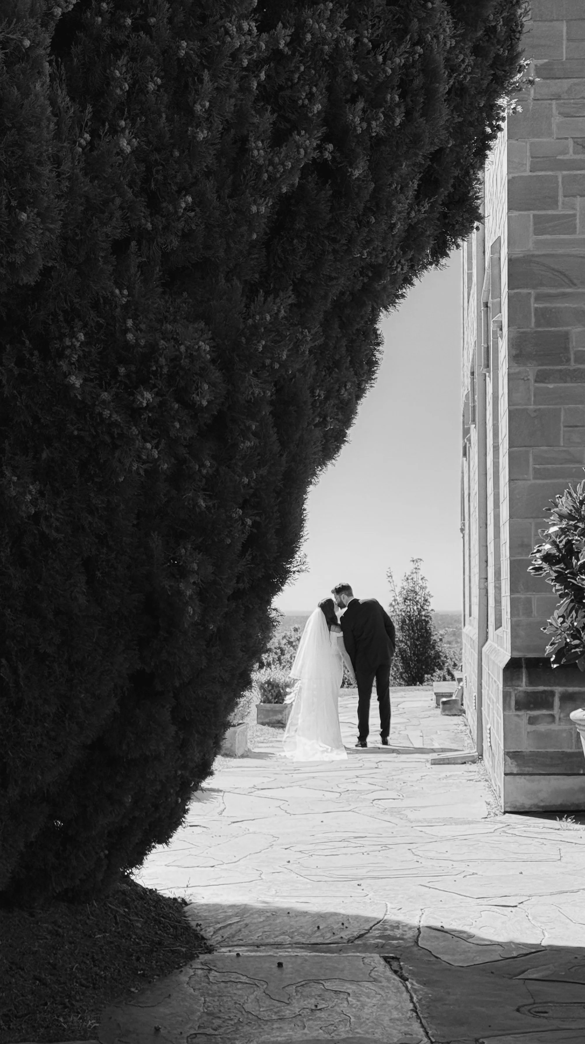 A bride and groom stand close together outdoors near a stone building, sharing a kiss or whisper, framed by large trees and a clear sky.