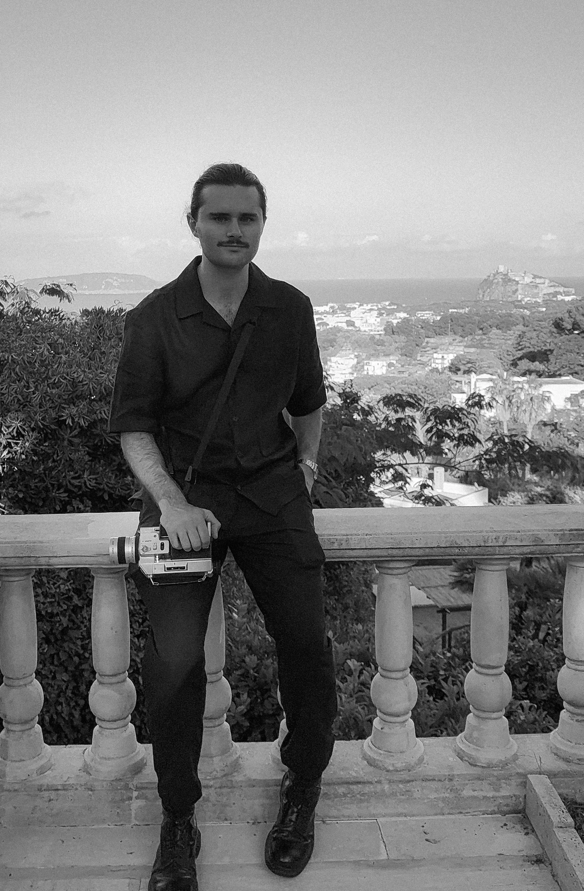 A man with a mustache and slicked-back hair standing outdoors in front of a scenic view with trees and buildings, holding a vintage camera, dressed in dark clothing, in black and white photo.
