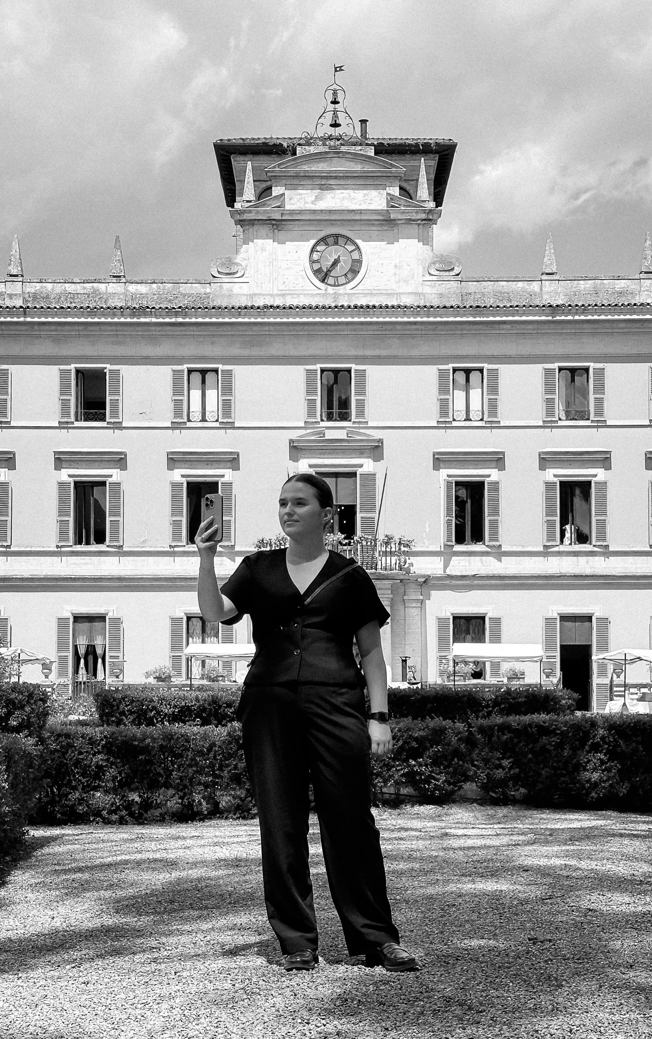 A woman is taking a selfie in front of a large historic building with a clock tower, multiple windows with shutters, and a flag on top, in black and white.