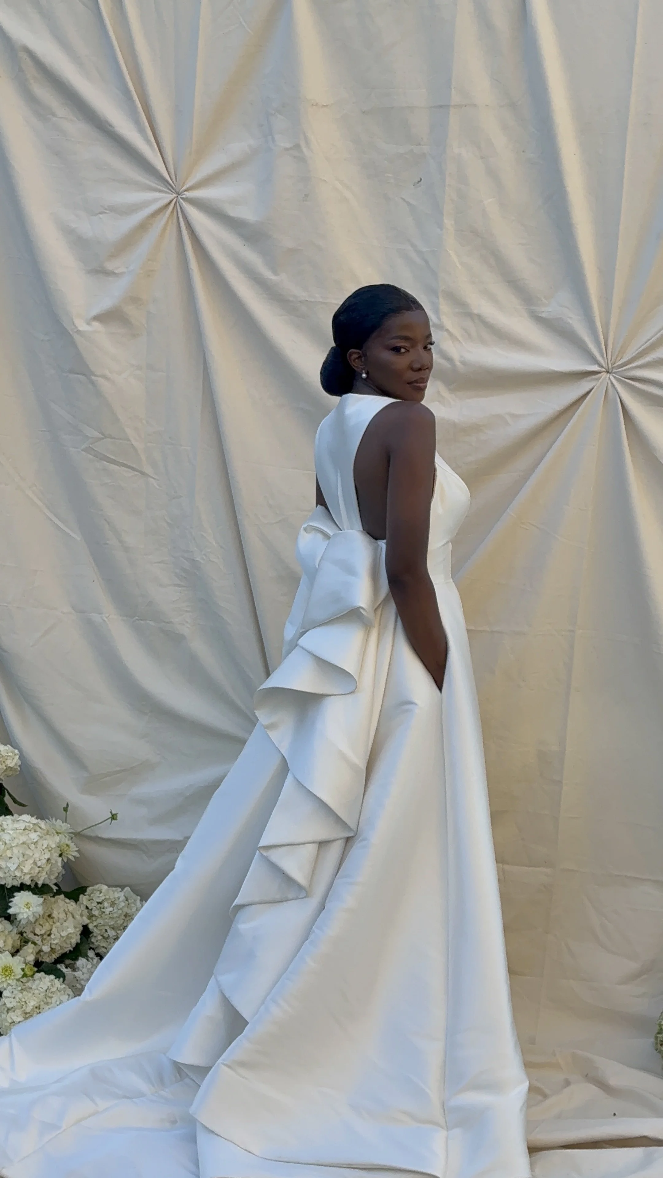A woman in a white, elegant dress with ruffles, standing against a cream-colored fabric backdrop, with white flowers on the floor nearby.
