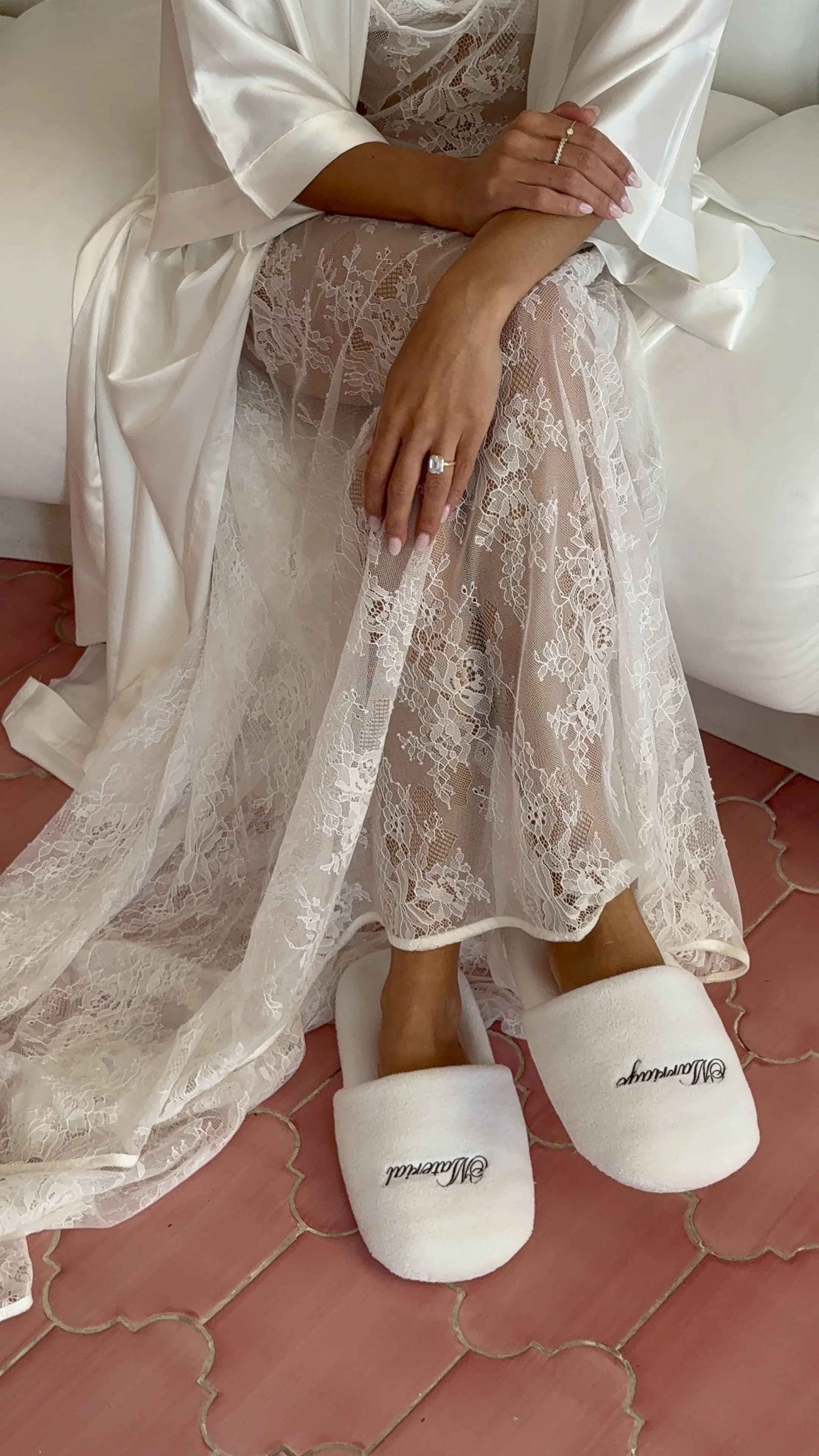 A woman in lace dress and white satin robe sitting on a couch, wearing fluffy white slippers with the word 'Bachata' embroidered on them, with hands resting on her lap showing rings.