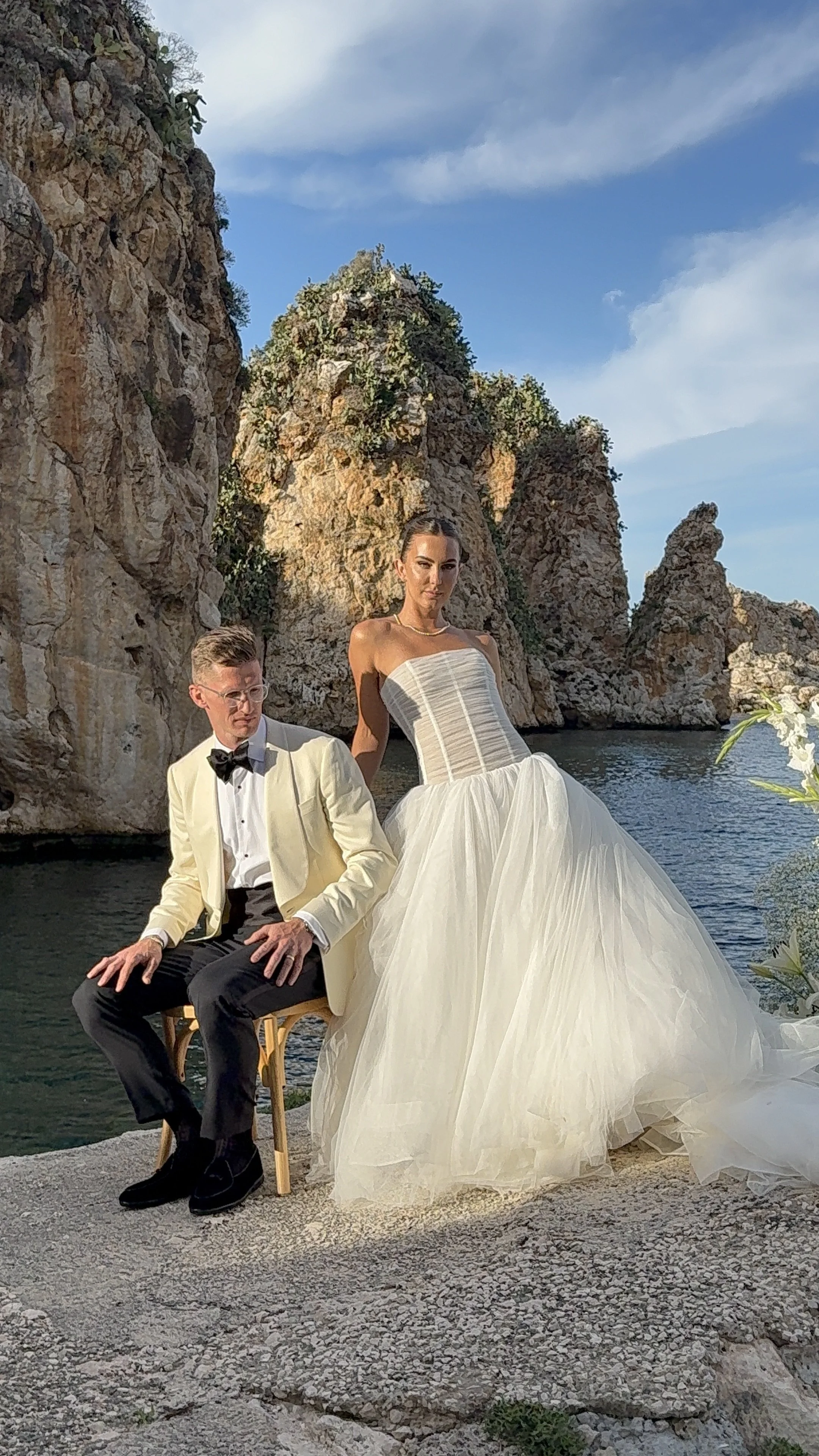 A bride and groom posing outdoors by the water on a rocky landscape at sunset. The groom is seated on a wooden chair wearing a white tuxedo jacket, black pants, and a bow tie. The bride is standing beside him in a strapless white wedding gown with a 