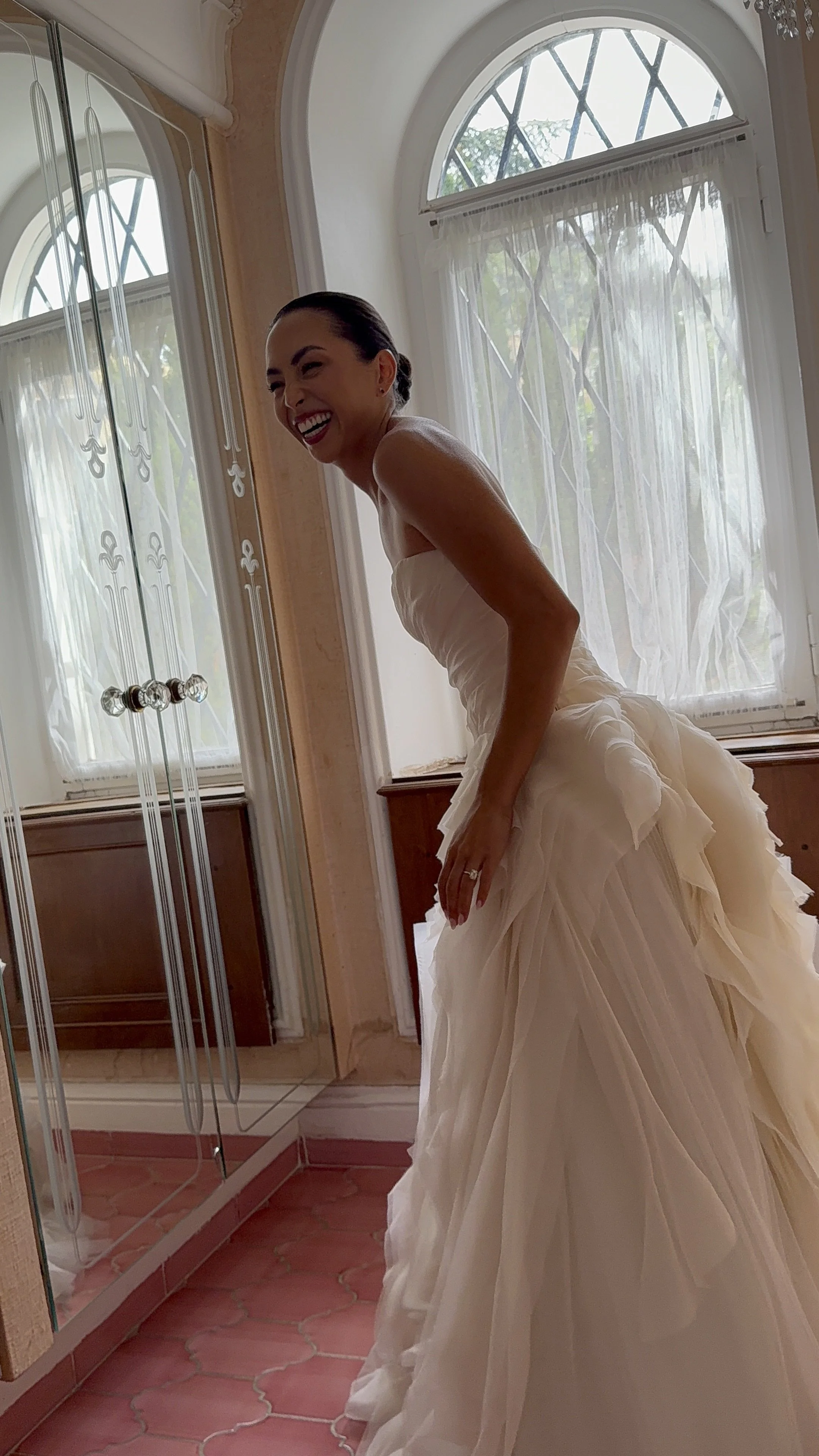 A smiling woman in a wedding dress standing in a room with large arched windows and a mirrored closet.