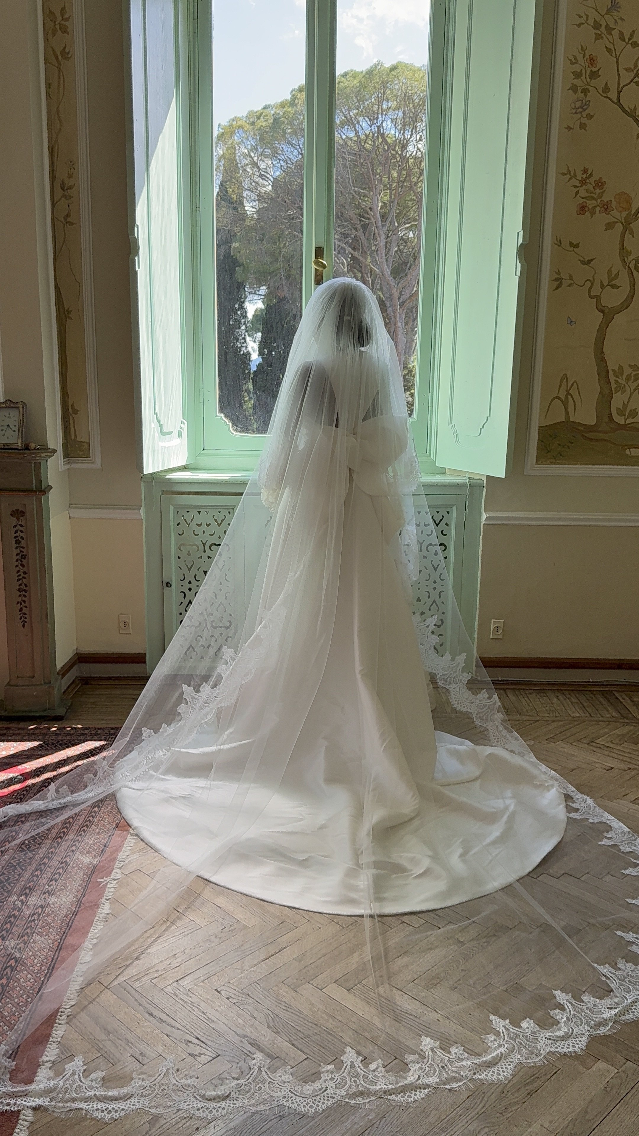 A bride in a white wedding dress with a long veil standing in front of an open window, looking outside at trees.