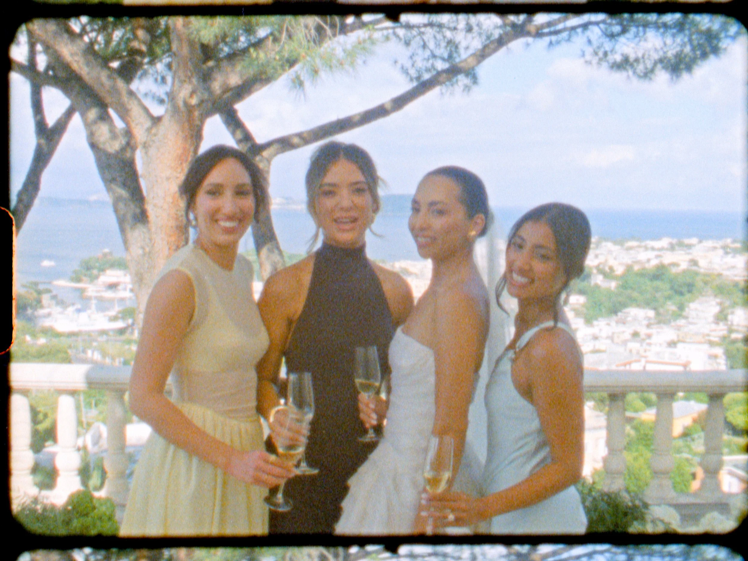 Four women in formal dresses holding champagne glasses, standing outdoors with trees and a cityscape background.
