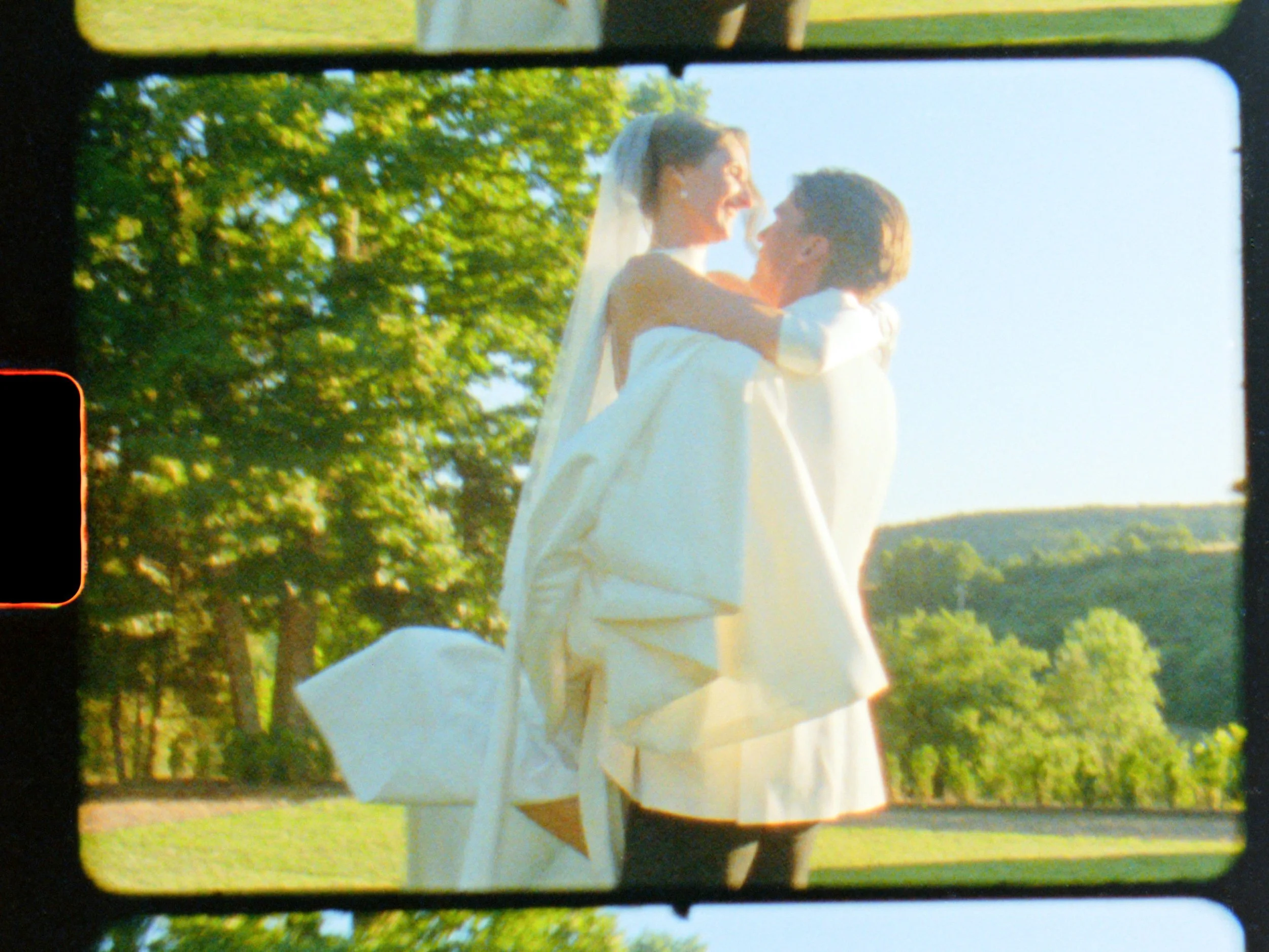 A bride and groom in wedding attire are outdoors, with the groom lifting the bride who has a veil and is smiling, set against a background of green trees and hills in the distance during daylight.