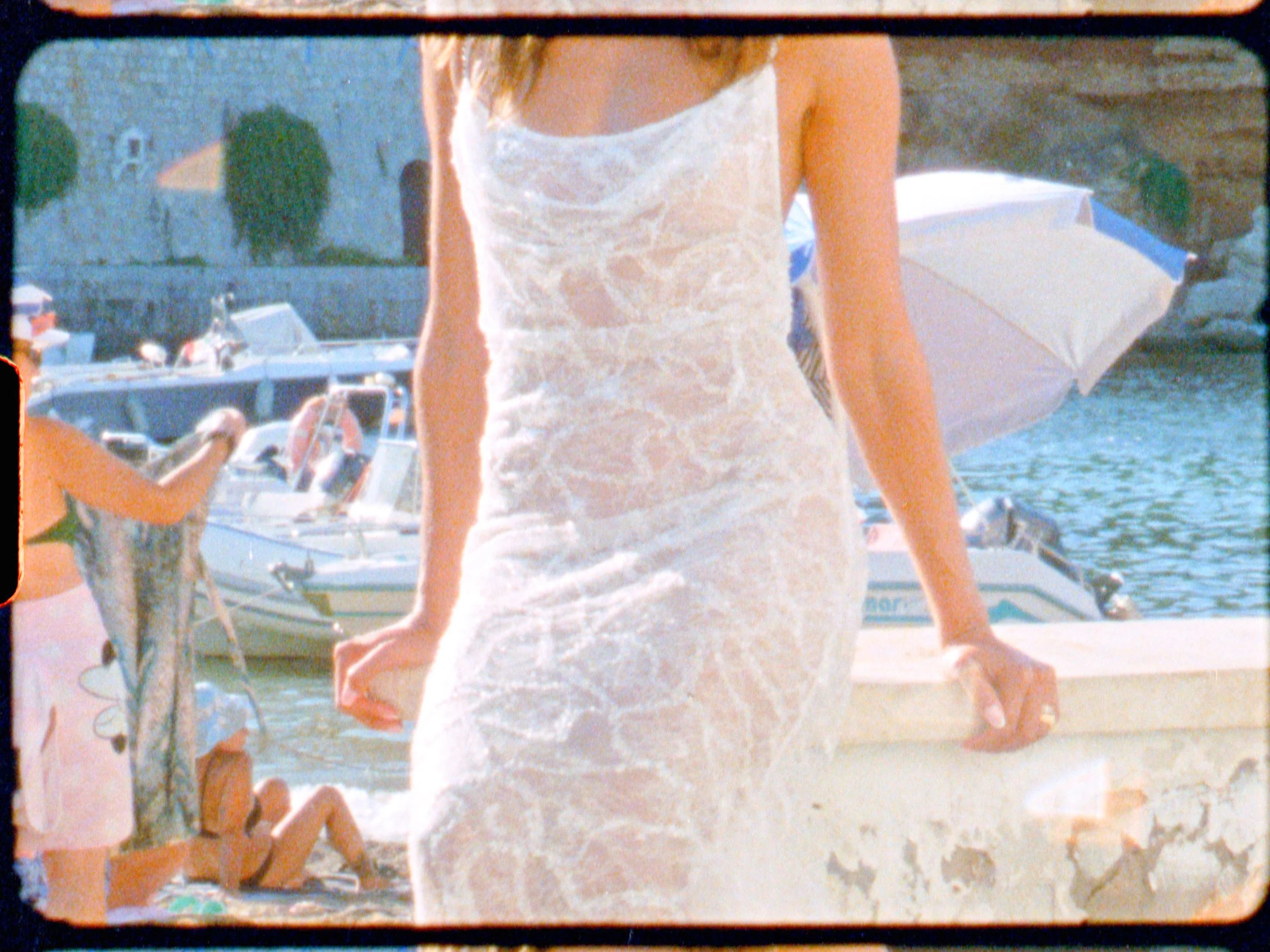 Woman in a white dress standing near a marina with boats, a young girl lying on the ground, and an umbrella in the background.