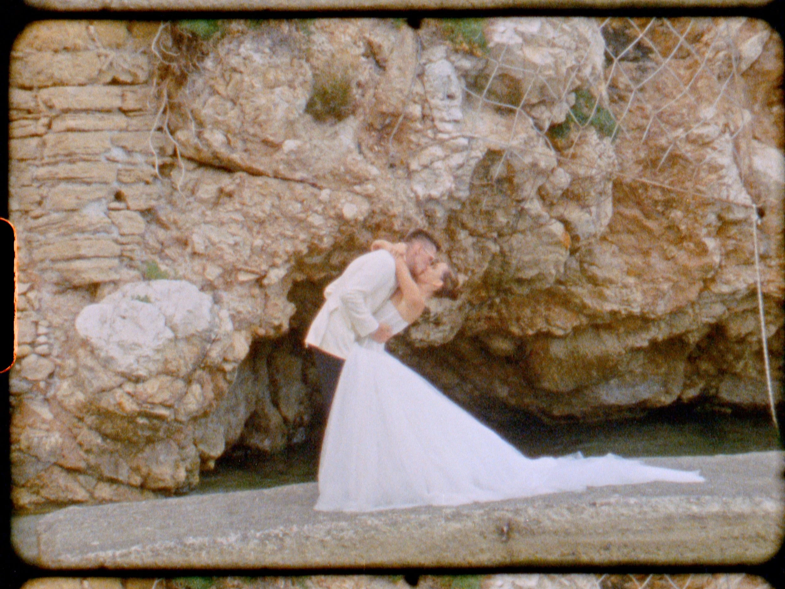 A woman dressed in a white gown and a man in formal attire are embracing underneath a rocky overhang on a beach.