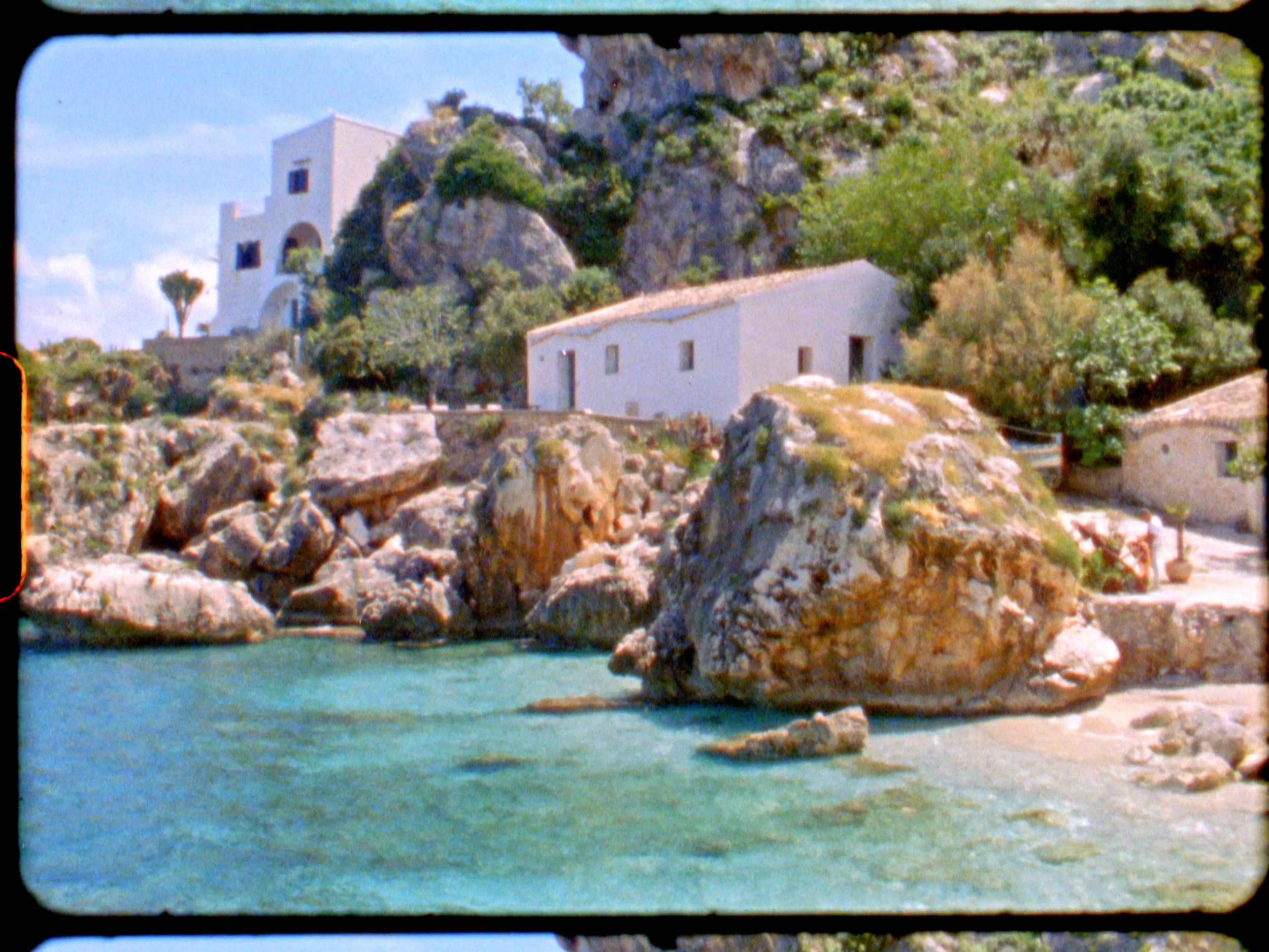 Cliffs with white buildings beside a turquoise sea, greenery on the rocky terrain.