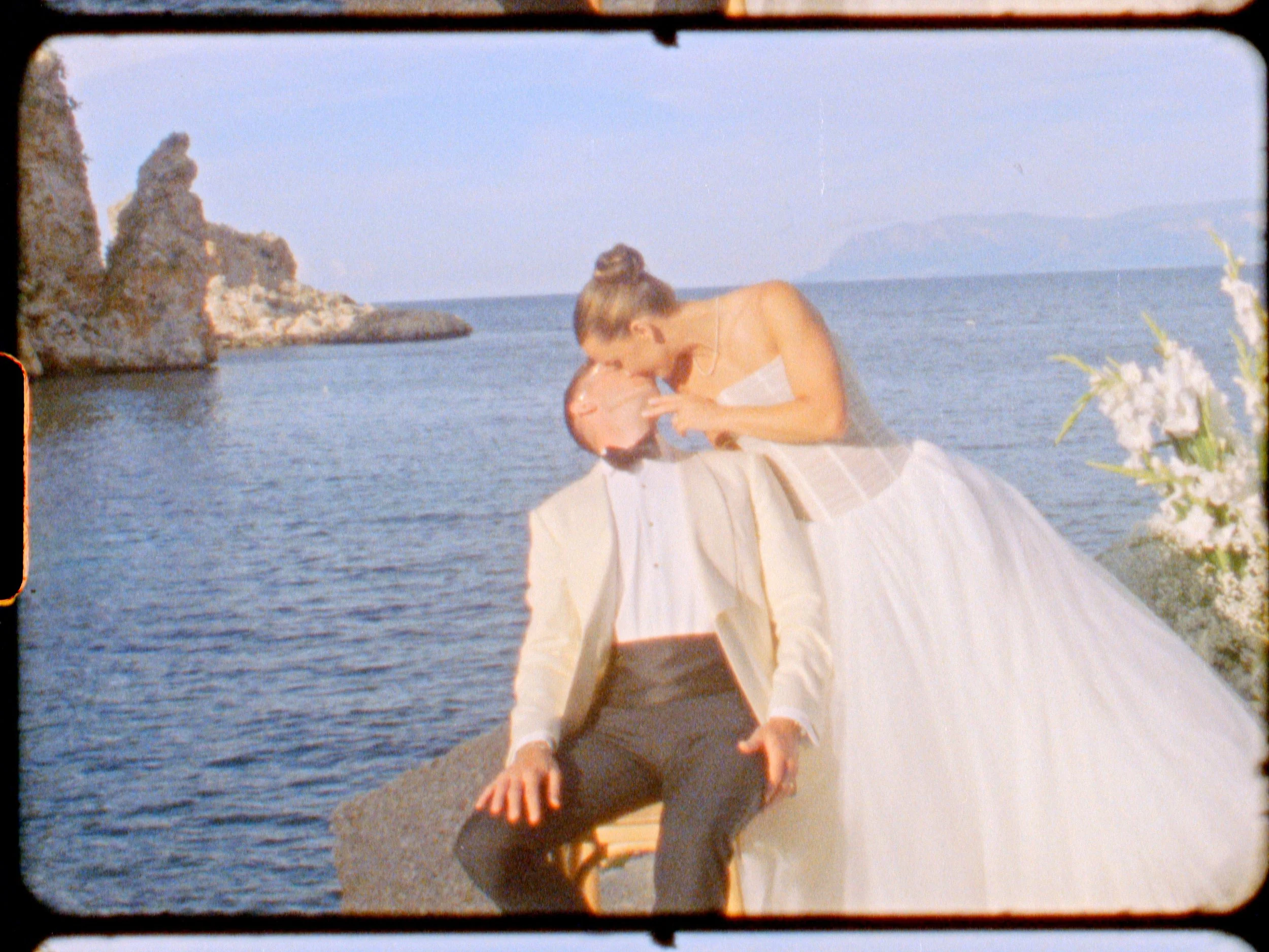 A woman in a wedding dress and a man in a suit by the water, with rocks and cliffs in the background.