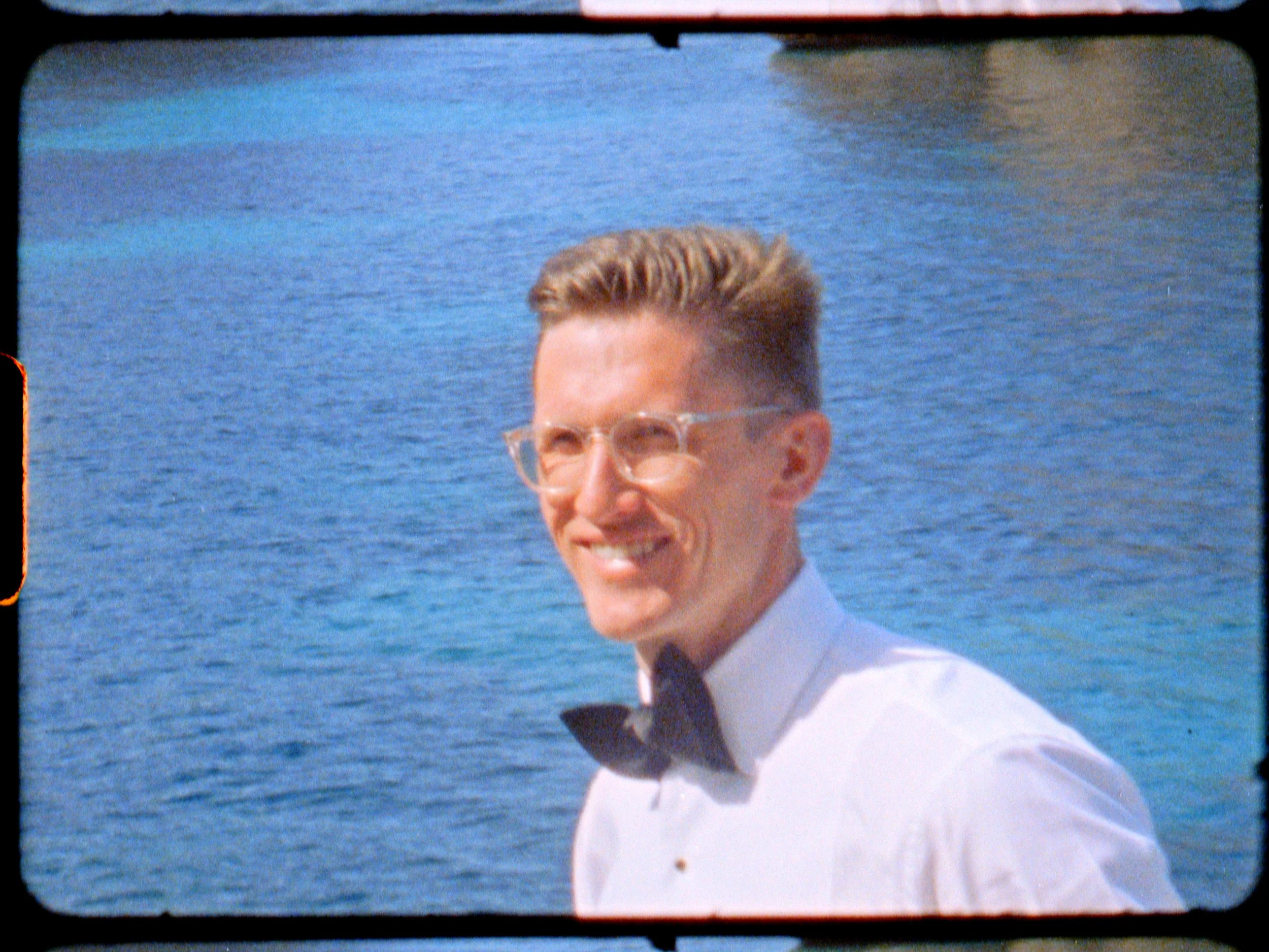 A smiling man with short, styled hair and clear glasses, wearing a white tuxedo shirt with a black bow tie, standing against a blue water background.