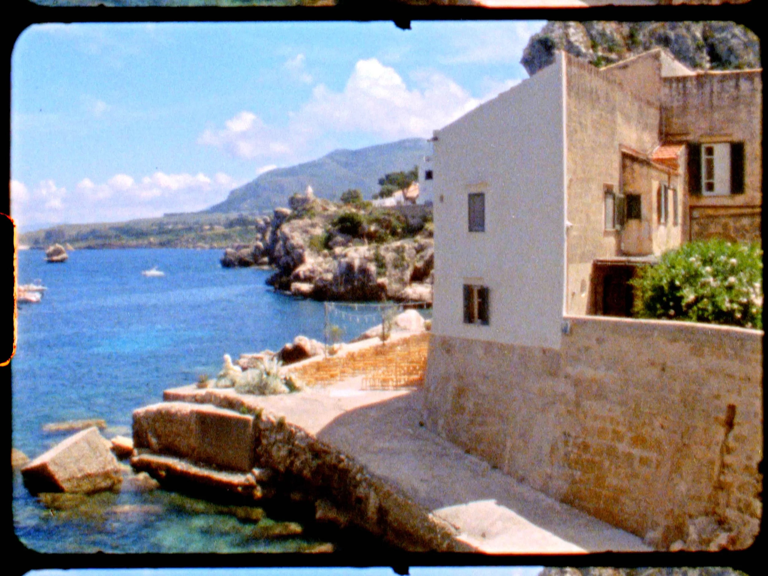 A coastal scene featuring a stone building with multiple windows, a stone wall, a pathway, and rocky shoreline along blue waters with boats and distant hills under a partly cloudy sky.