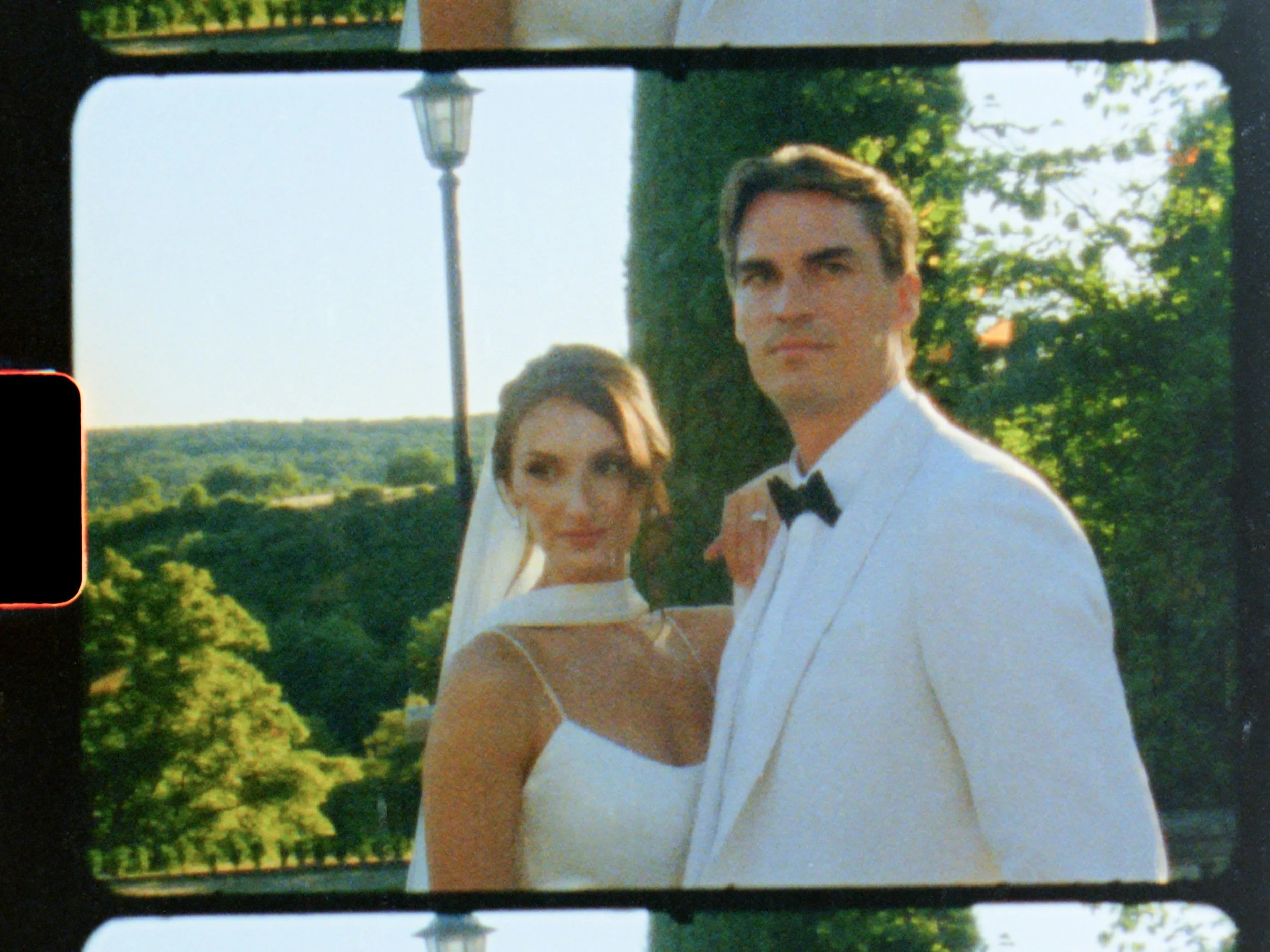 A bride and groom standing outdoors during daytime, with trees and hills in the background. The bride is wearing a white dress and veil, and the groom is dressed in a white tuxedo with a black bow tie.