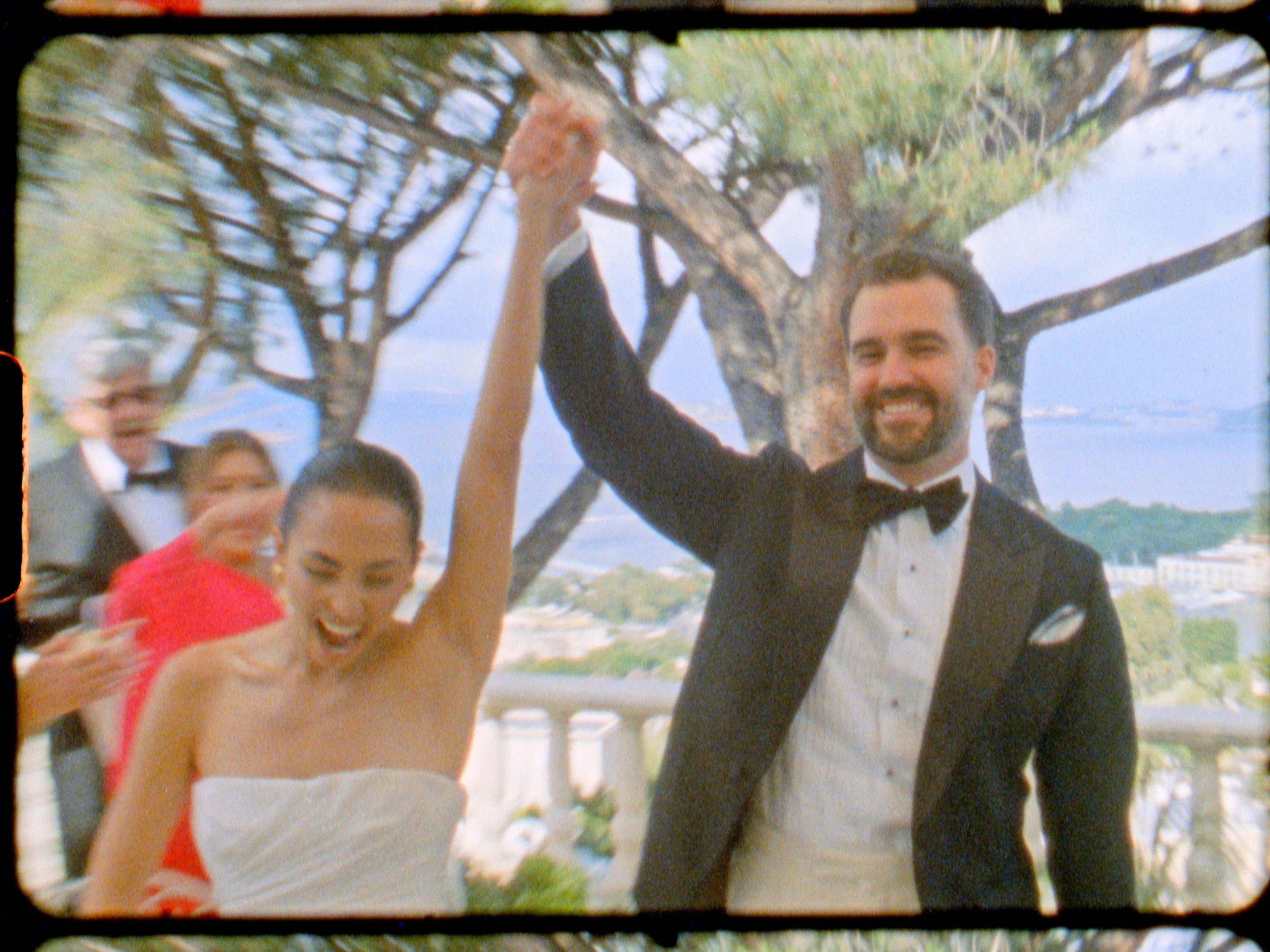 A happy couple, the woman in a strapless white dress and the man in a tuxedo, celebrating outdoors near a tree with a scenic background, possibly at a wedding.