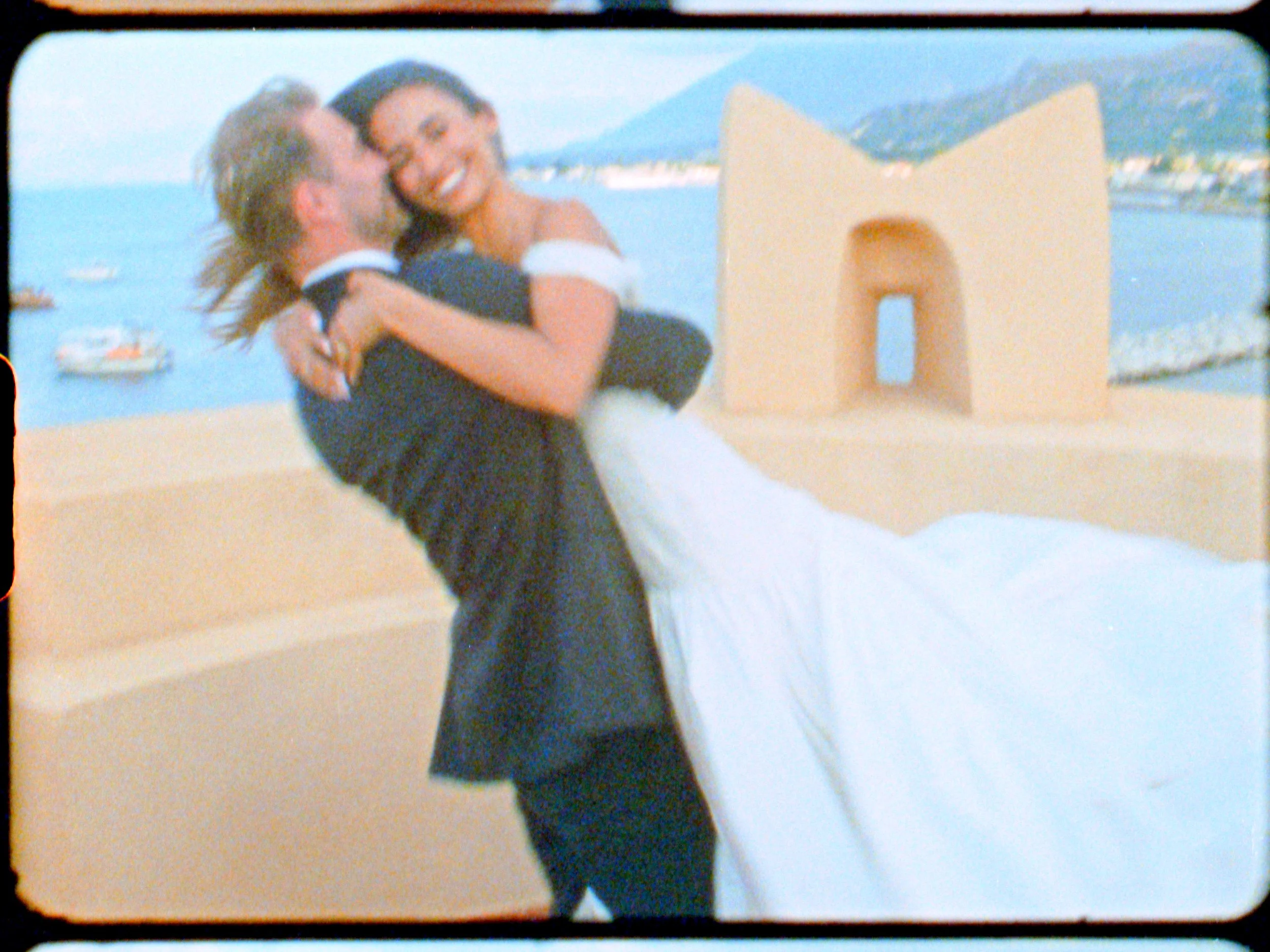 A bride and groom dancing outdoors near a body of water with boats and mountains in the background, and an architectural structure with an archway behind them.