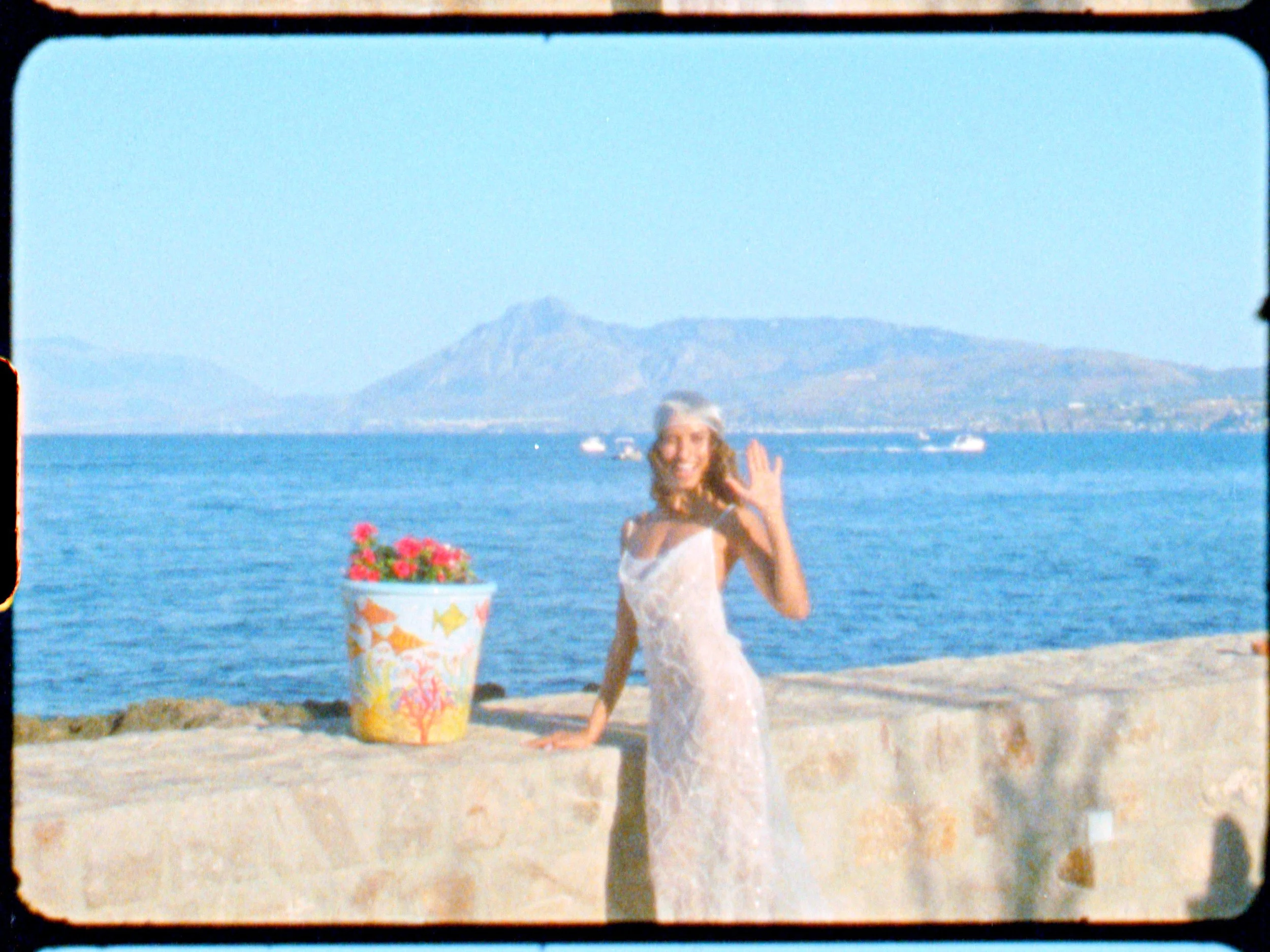 A woman in a strapless dress standing near a stone wall by the ocean, waving at the camera with a mountain and boats in the background.