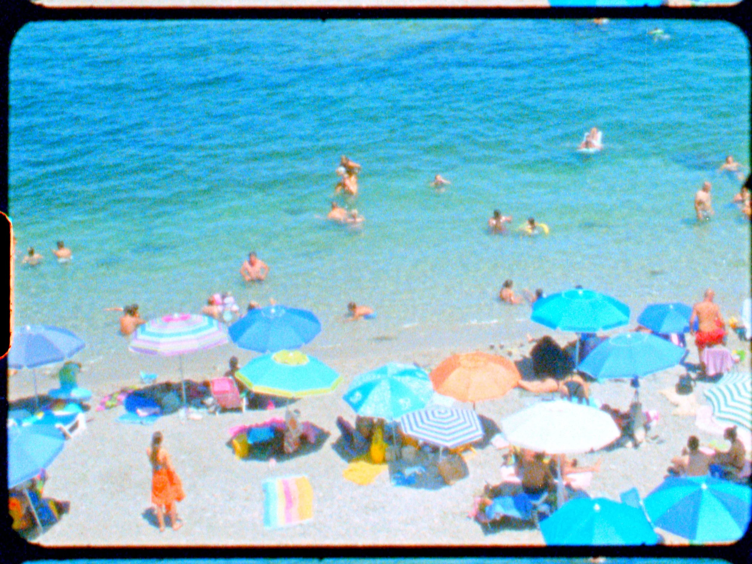 Crowded beach with colorful umbrellas, people swimming and relaxing near the shoreline, and a building with a red roof in the background.