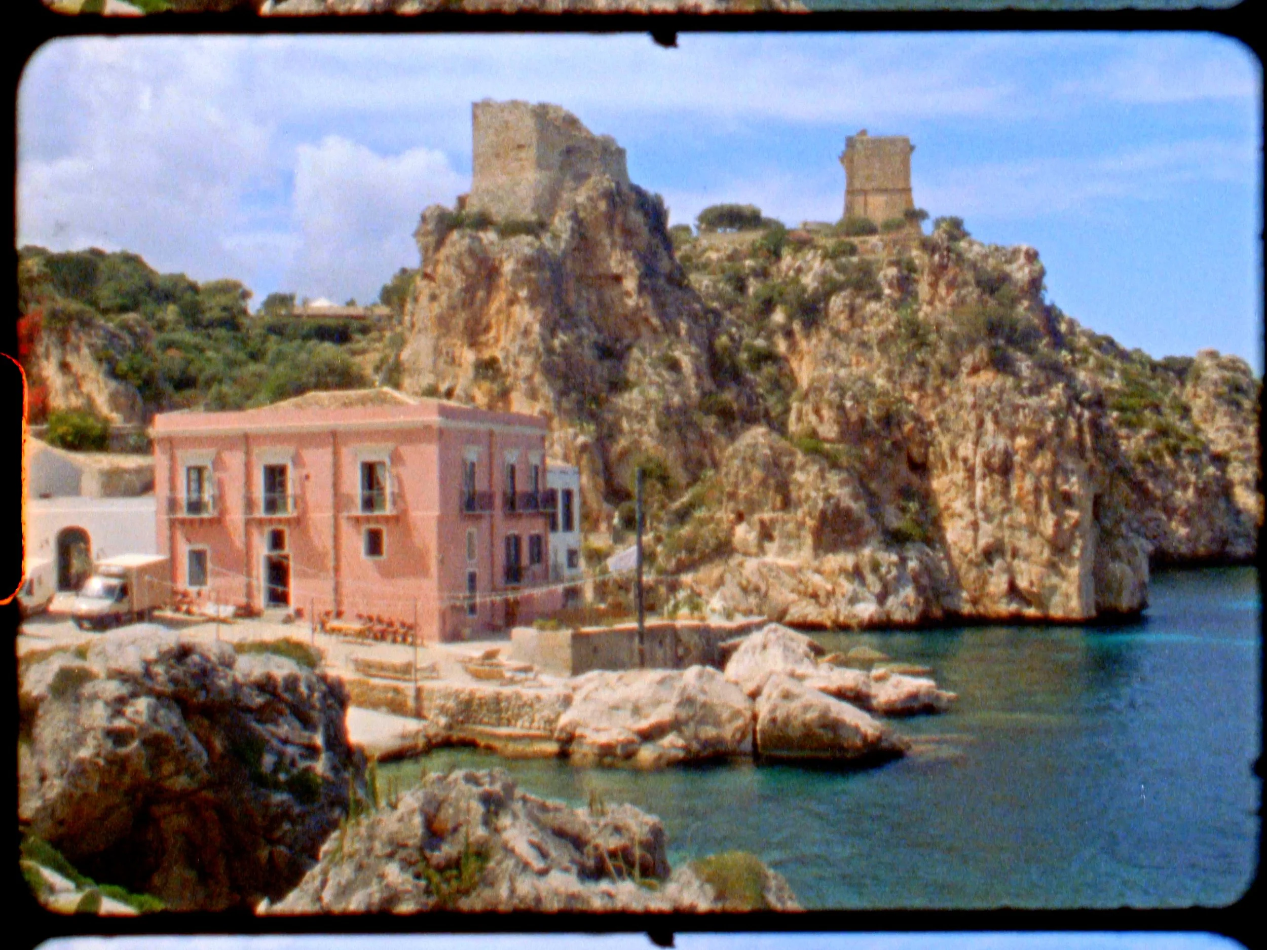 Pink building by the water with rocky cliffs and ruins on top of a hill in the background.