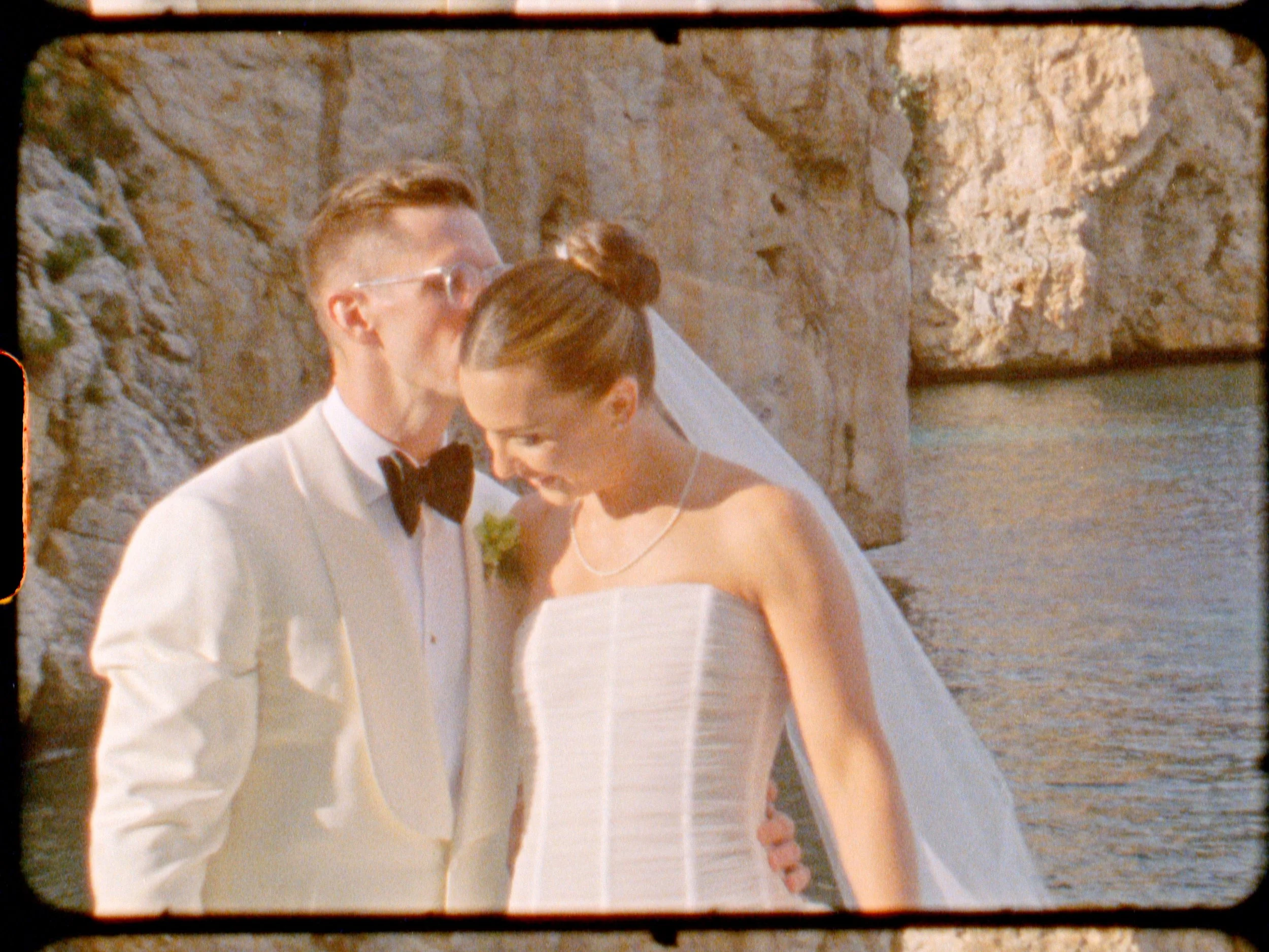 A newlywed couple dressed in wedding attire stands close together near rocky cliffs and water, with the groom wearing a cream tuxedo and the bride in a strapless white gown and veil.