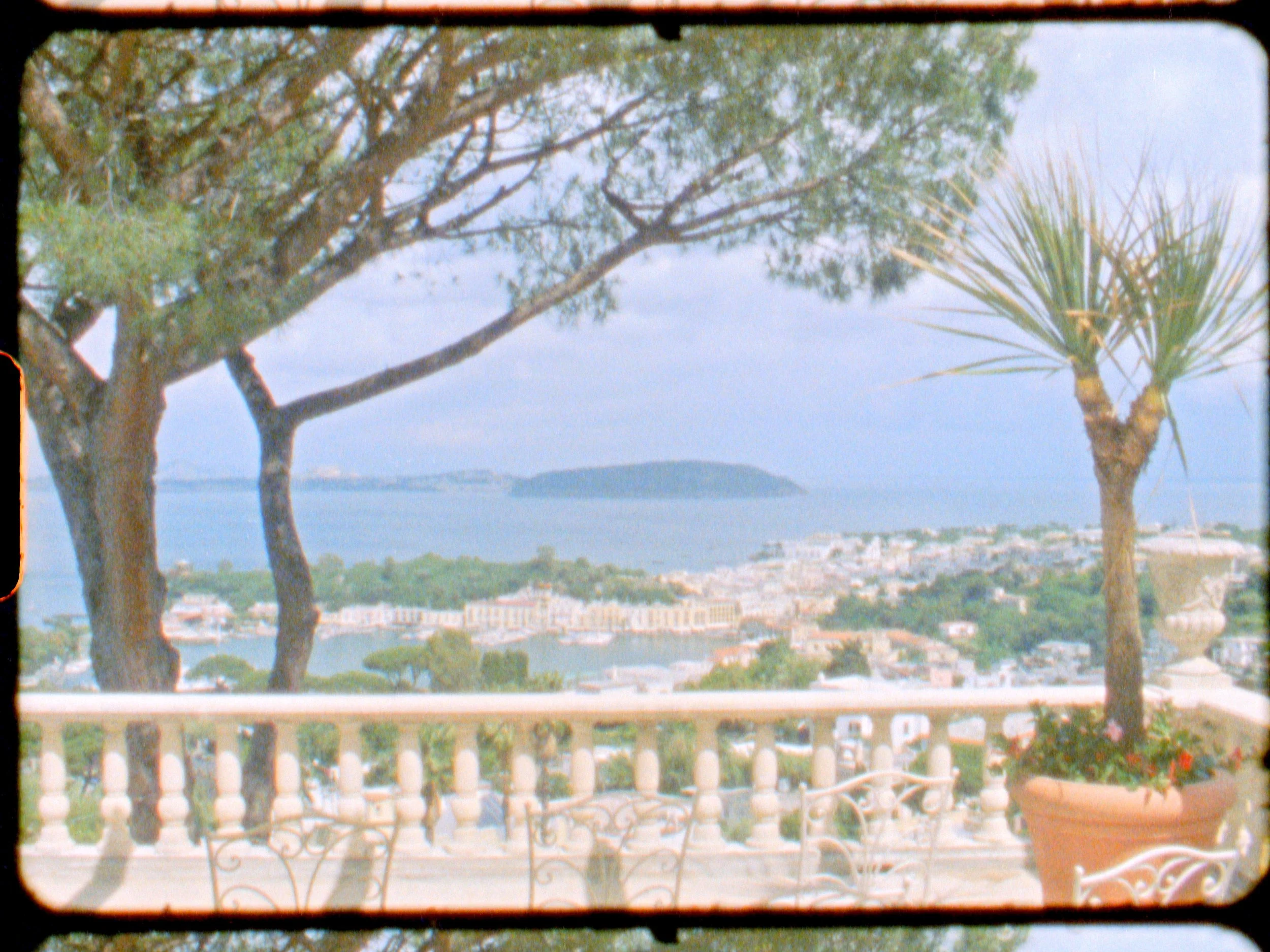 View of a coastal town from a balcony with trees, plants, and decorative chairs.