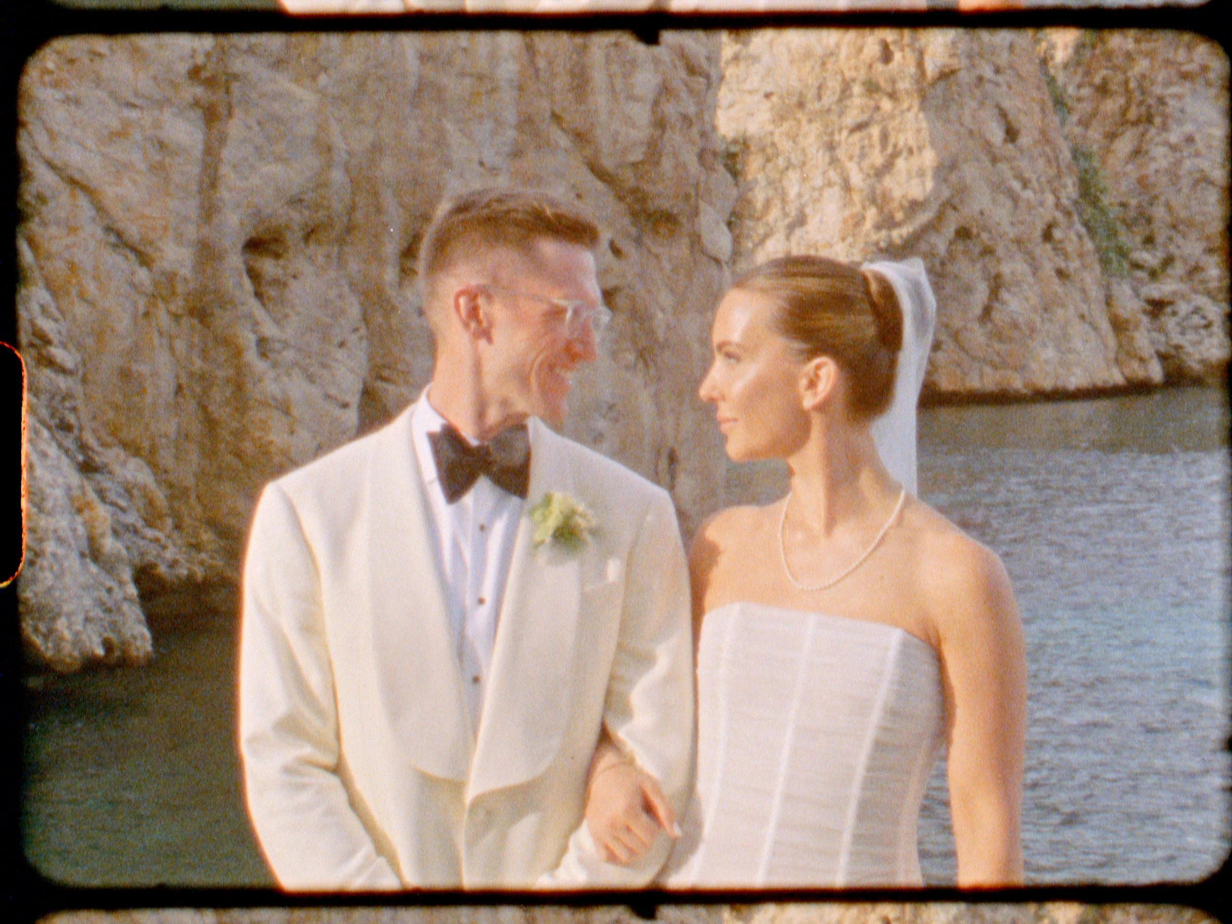 A man and woman dressed in wedding attire, standing by rocky coastline with water in the background, smiling at each other.