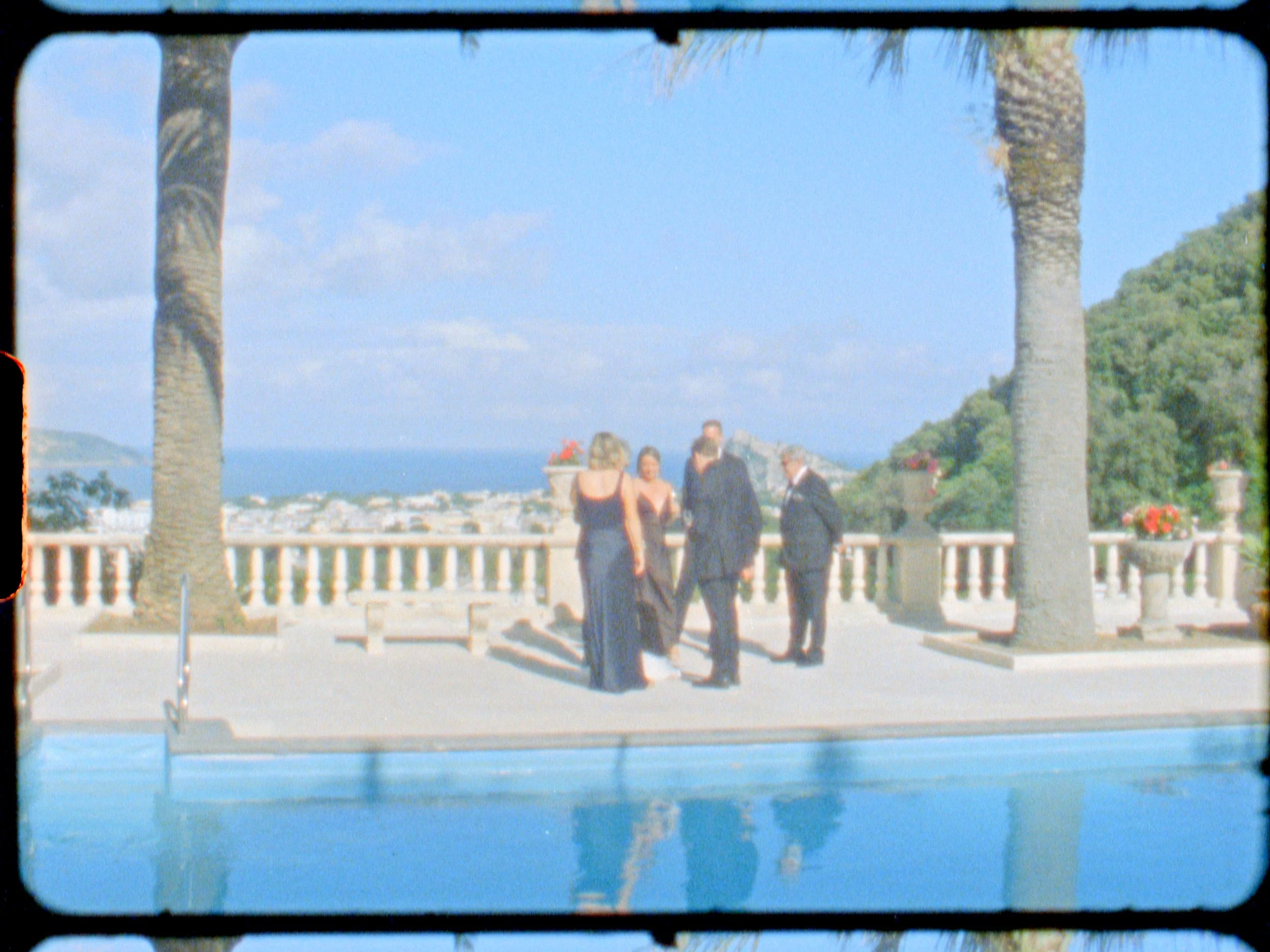 People in formal attire standing on a terrace by a pool, with a scenic coastal view and palm trees in the background.