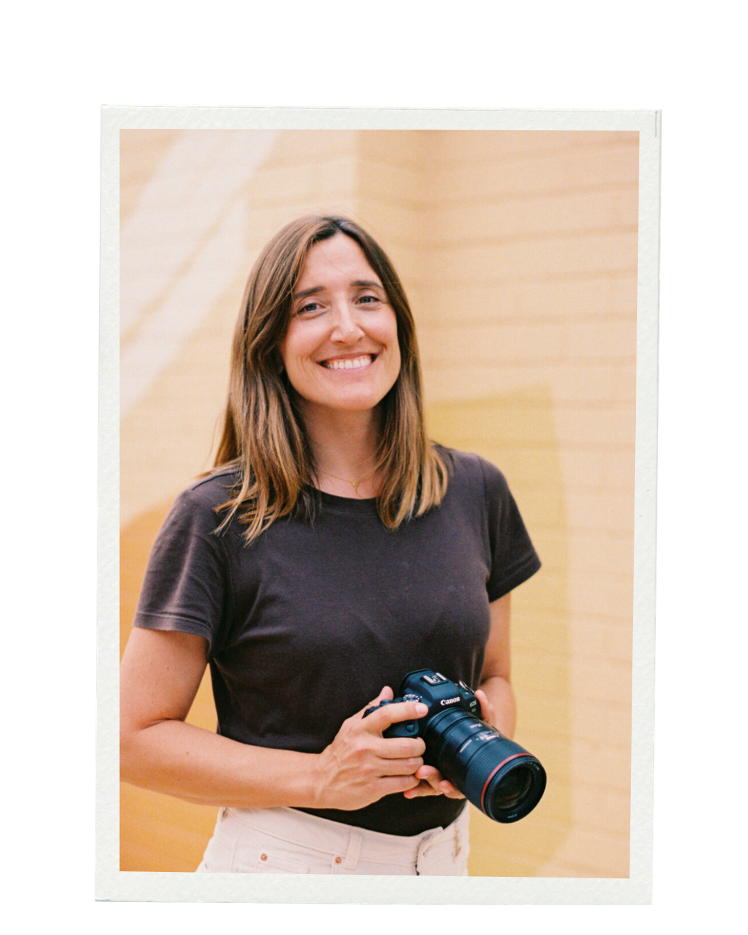 A smiling woman with shoulder-length brown hair holding a Canon camera, standing in front of a light beige background. Branding & Websites Northern Rivers NSW. Elin Bandmann Photography.