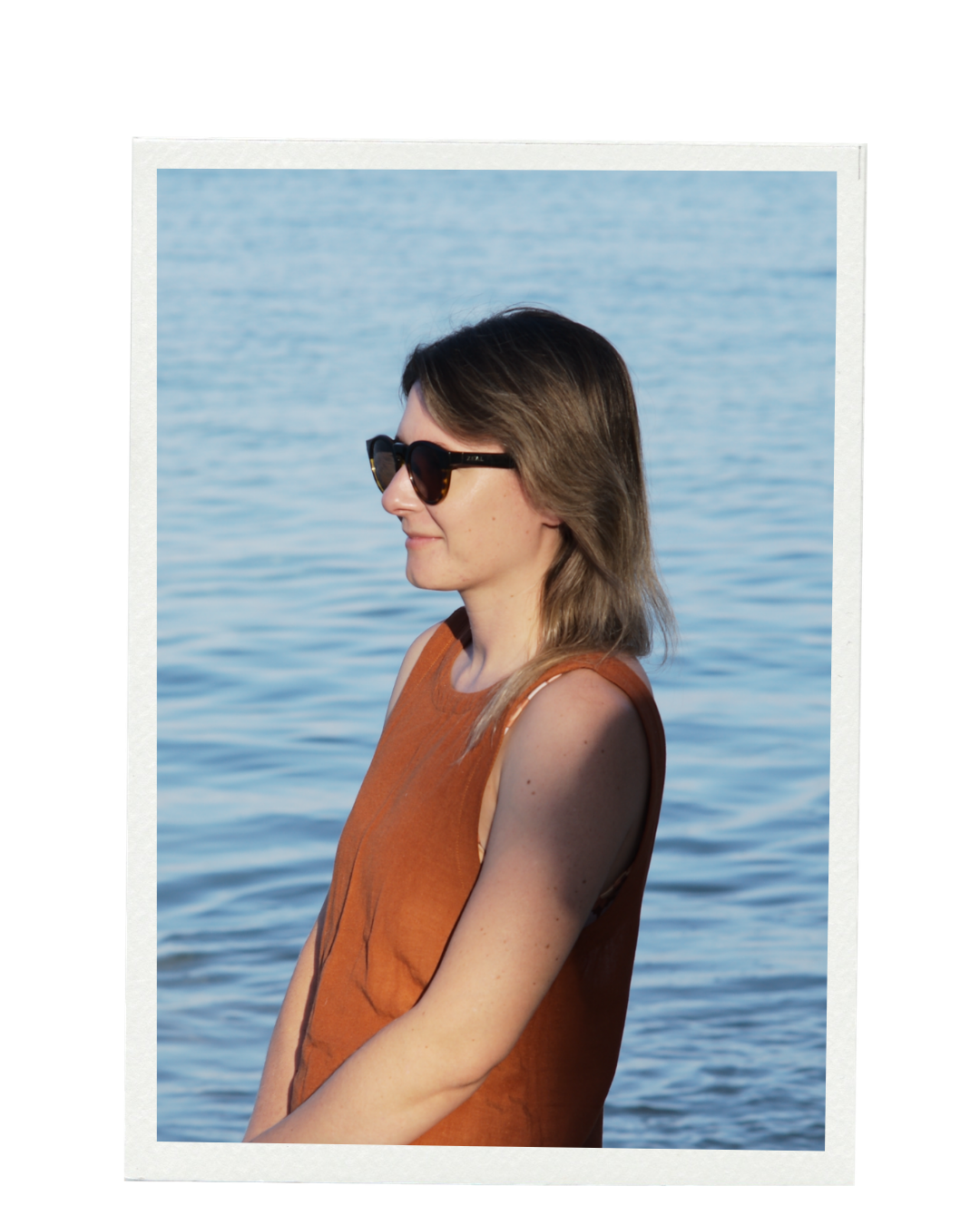 A woman with shoulder-length brown hair and sunglasses standing near a body of water, wearing an orange sleeveless top. Coral Theory. Branding & Websites Northern Rivers NSW
