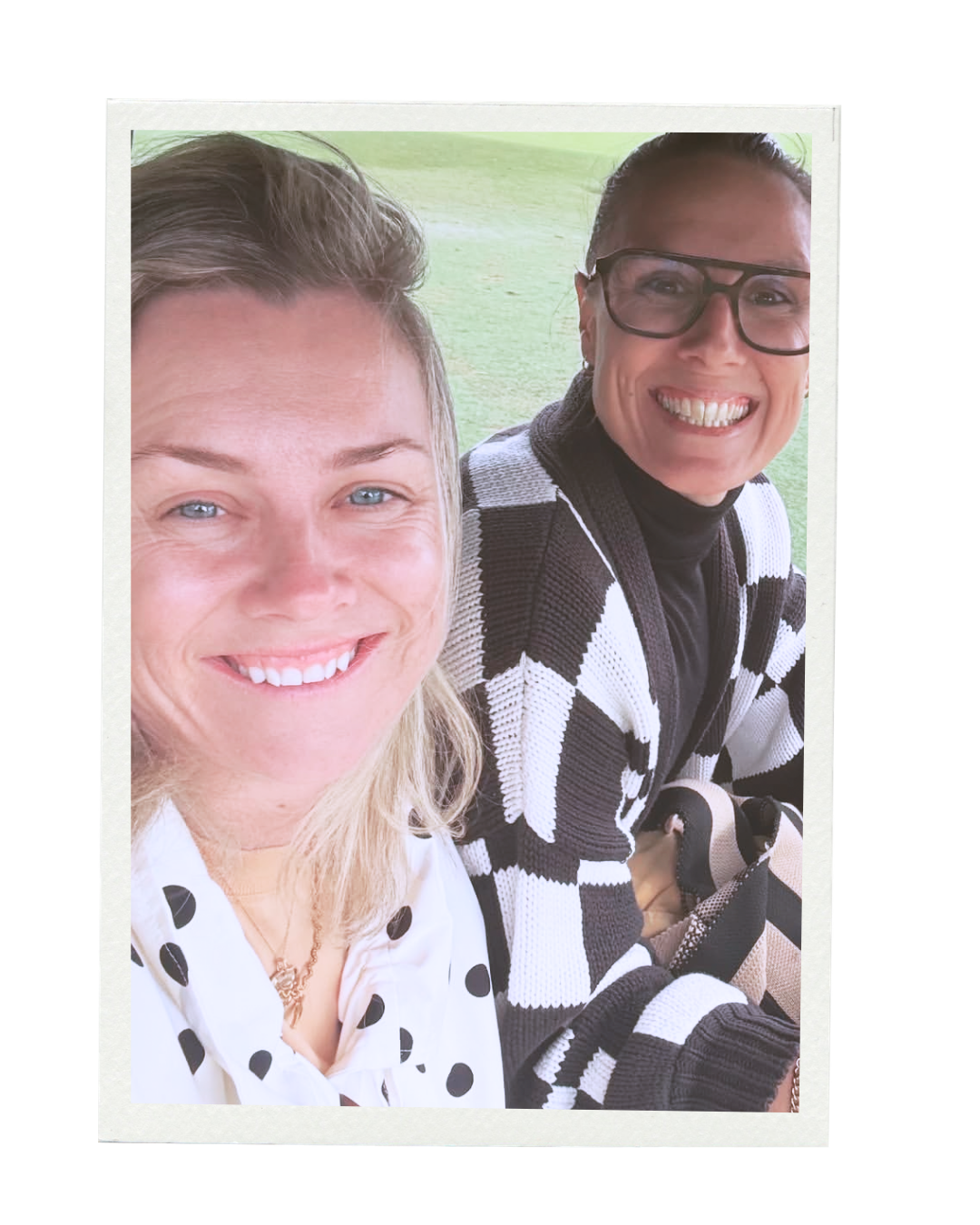 Two smiling women taking a selfie outdoors on a grassy area. The woman on the left has blonde hair, blue eyes, and is wearing a white shirt with black polka dots. The woman on the right has dark hair, glasses, and is wearing a black and white patterned sweater.