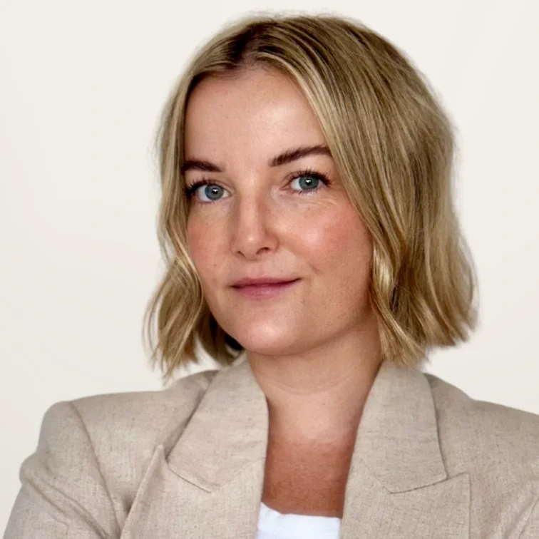 A woman with shoulder-length blonde hair, blue eyes, and light skin, wearing a beige blazer and white top, looking directly at the camera with a slight smile.