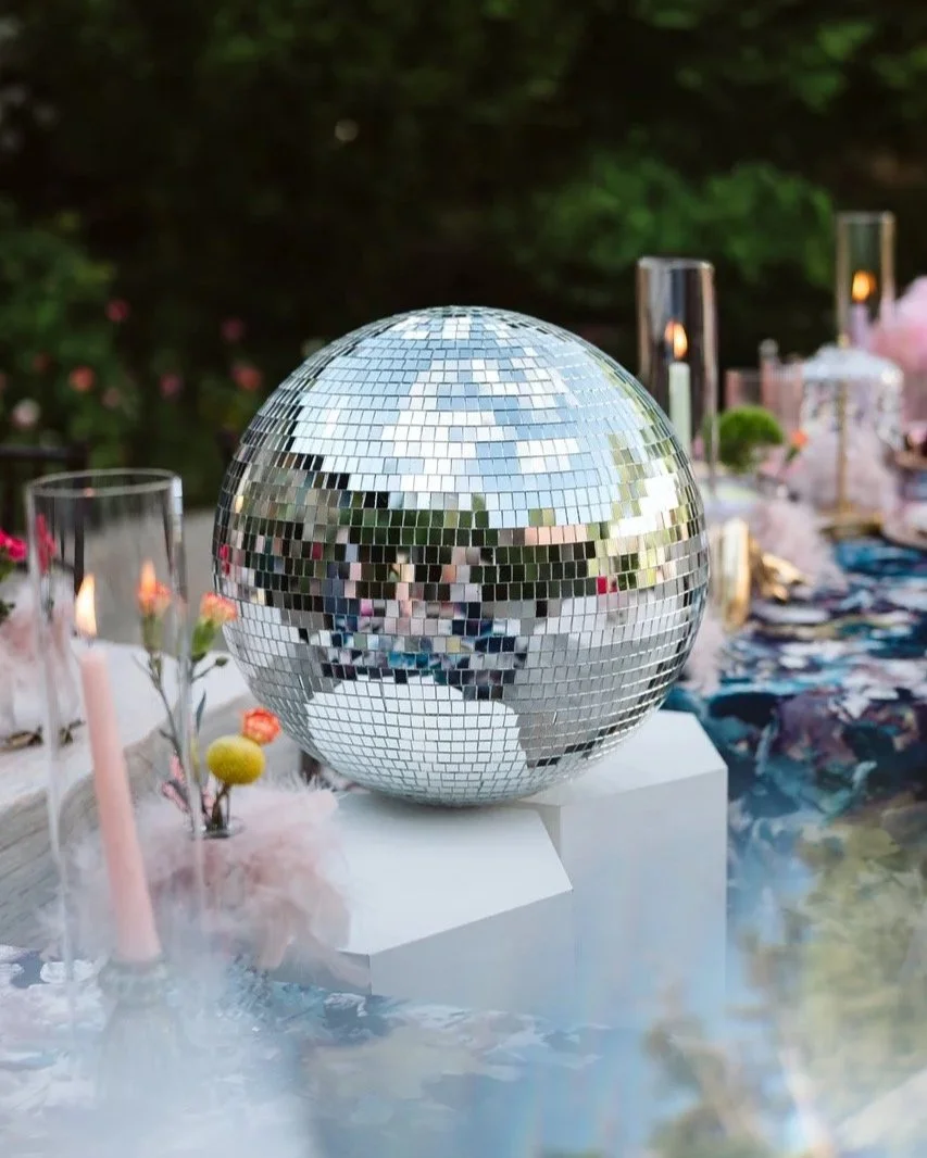 A large mirror disco ball on a white stand at an outdoor party table.