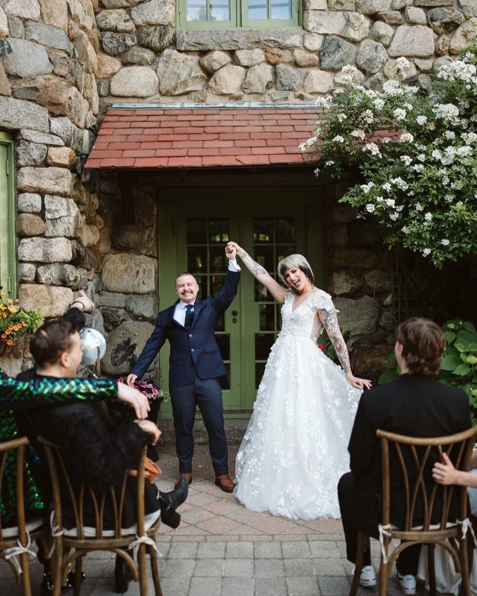 A newlywed couple celebrates at their wedding reception outside a stone building. The groom in a navy suit raises the bride's hand, who is wearing a white wedding gown with lace details. Guests sit on wooden chairs, celebrating and smiling.