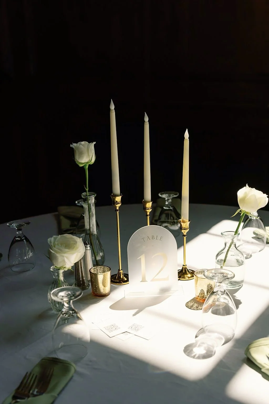 Elegant table arrangement with white roses in glass vases, three tall white candles in gold holders, a table number card labeled 'Table 12', and glassware on a white tablecloth, with sunlight highlighting the setup.