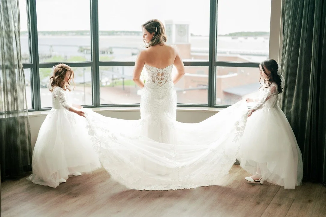 A bride in a white lace wedding gown standing by a window with two young girls in white dresses holding her train.