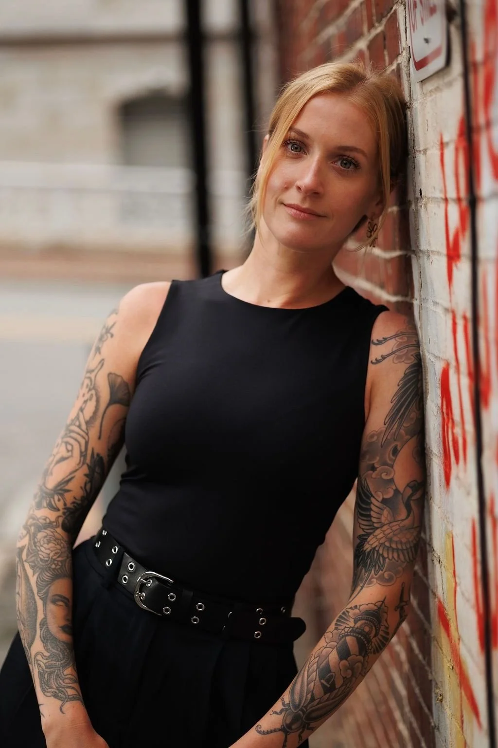 A woman with blonde hair and tattoos on her arms leaning against a graffiti-covered brick wall, wearing a black sleeveless top and black pants.