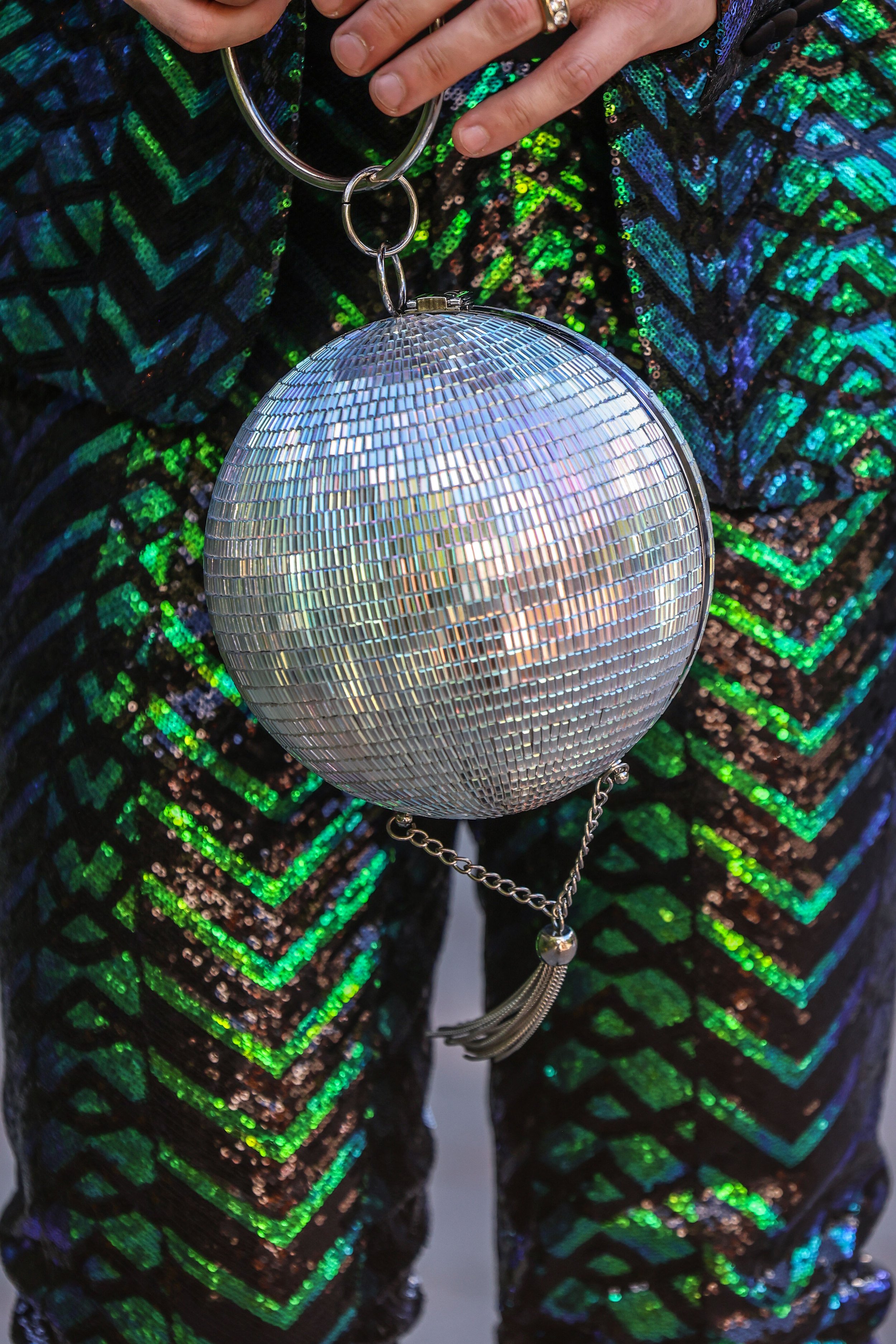 Close-up of a person's hand holding a shiny, disco ball purse with a chain strap, wearing a multicolored, sequined garment with a geometric pattern of green, blue, and black.