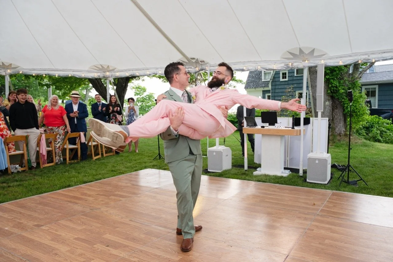 Groom in a light gray suit dancing with a man in a pink suit at an outdoor wedding reception, guests watching and smiling in the background under a tent.