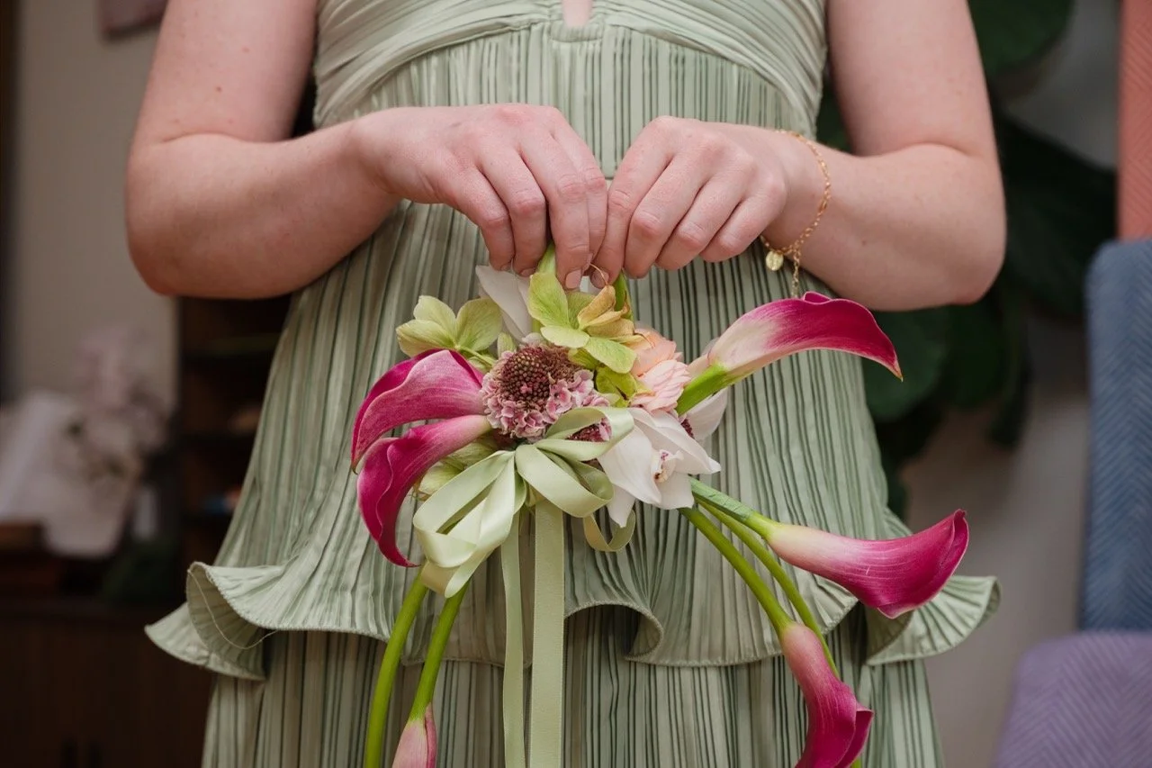 Person in a light green, ruffled dress holding a bouquet of pink, white, and green flowers, including calla lilies and other mixed blooms.
