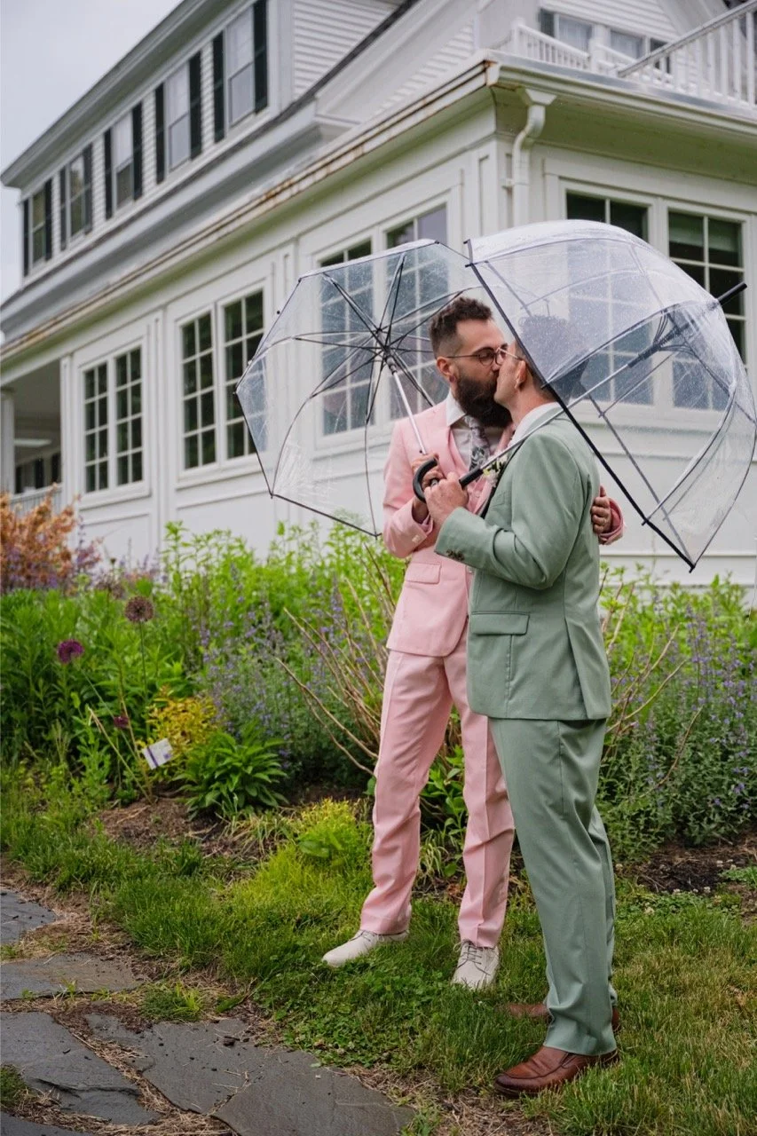 Two men dressed in pastel suits, one pink and one green, sharing a kiss under clear umbrellas in a garden in front of a white house.