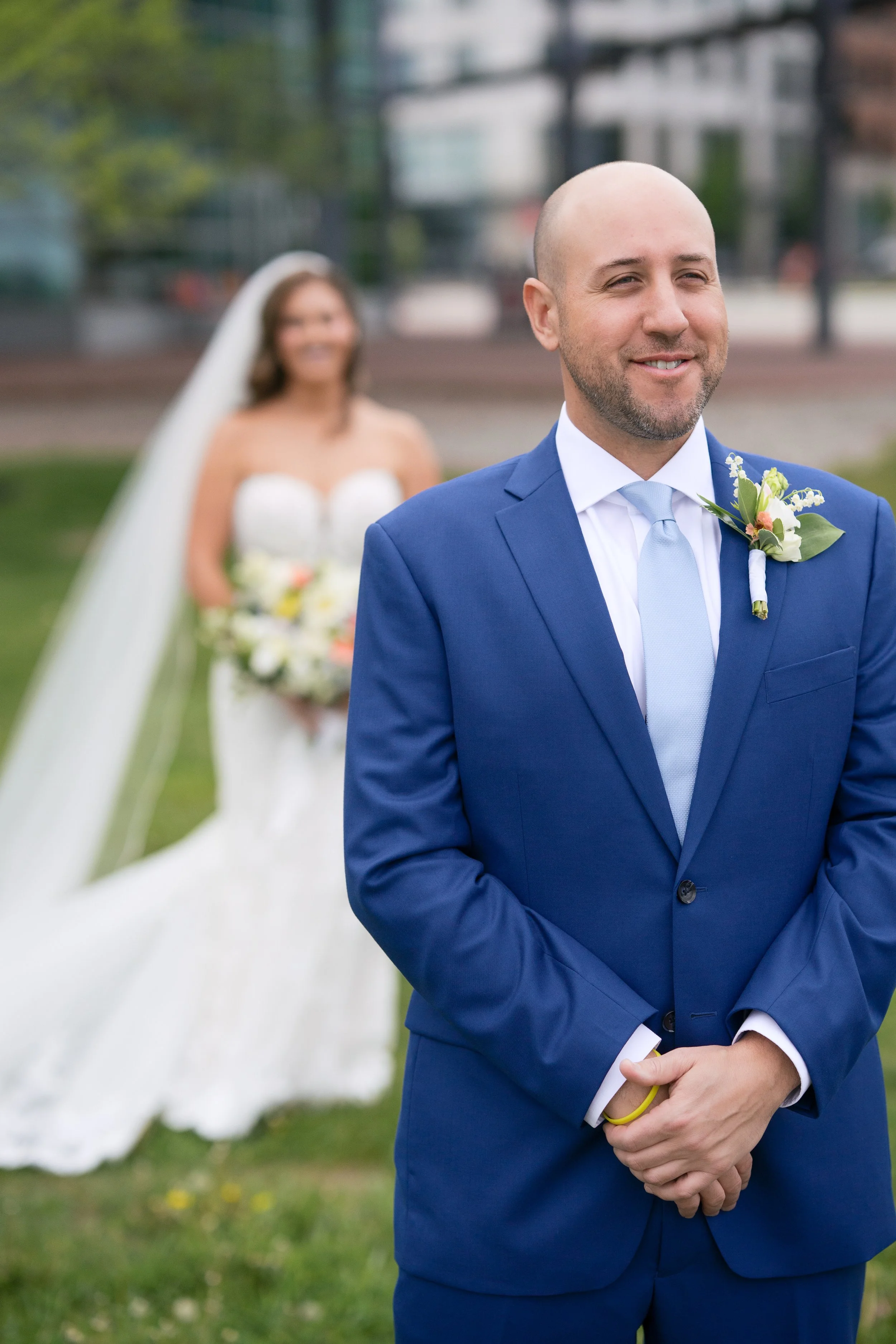 A man in a blue suit with a boutonnière smiling in the foreground, with a woman in a wedding dress and veil holding a bouquet blurred in the background during an outdoor wedding.