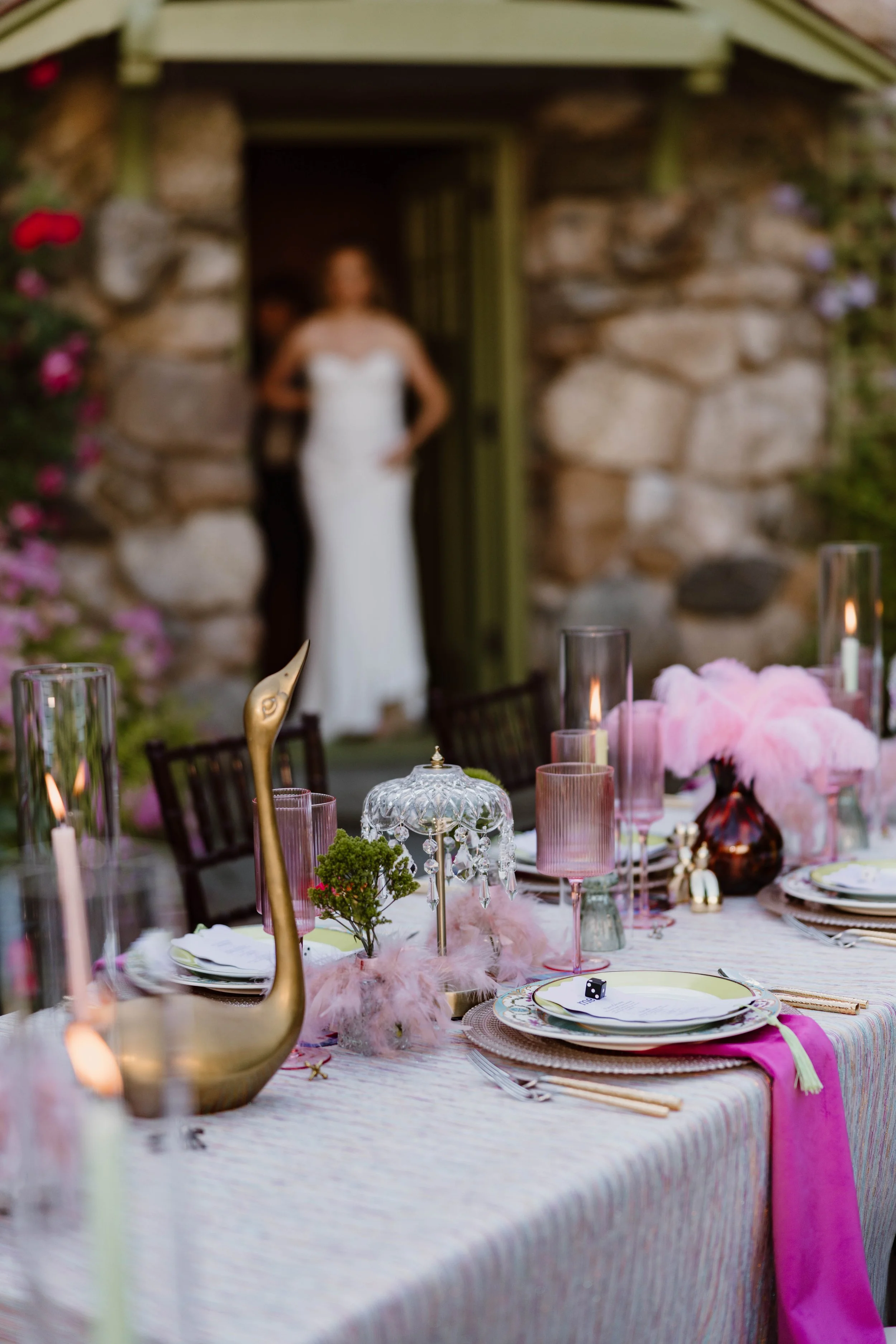 Blurred image of a woman in a white dress standing in a rustic stone building, with a decorated banquet table in the foreground featuring candles, flowers, and elegant tableware.