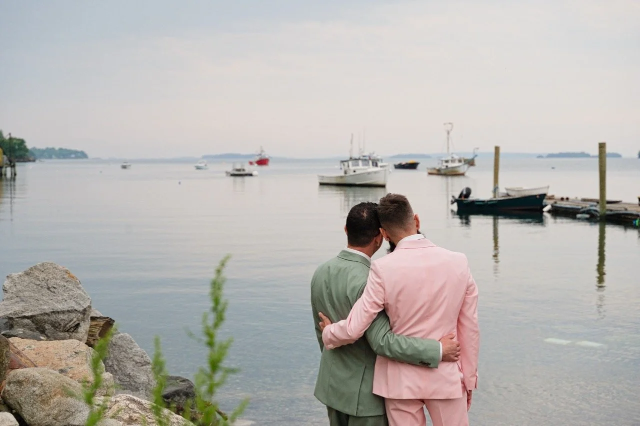 Two men dressed in pastel suits sharing an intimate moment by the water on a dock, with sailboats and yachts anchored in the background.