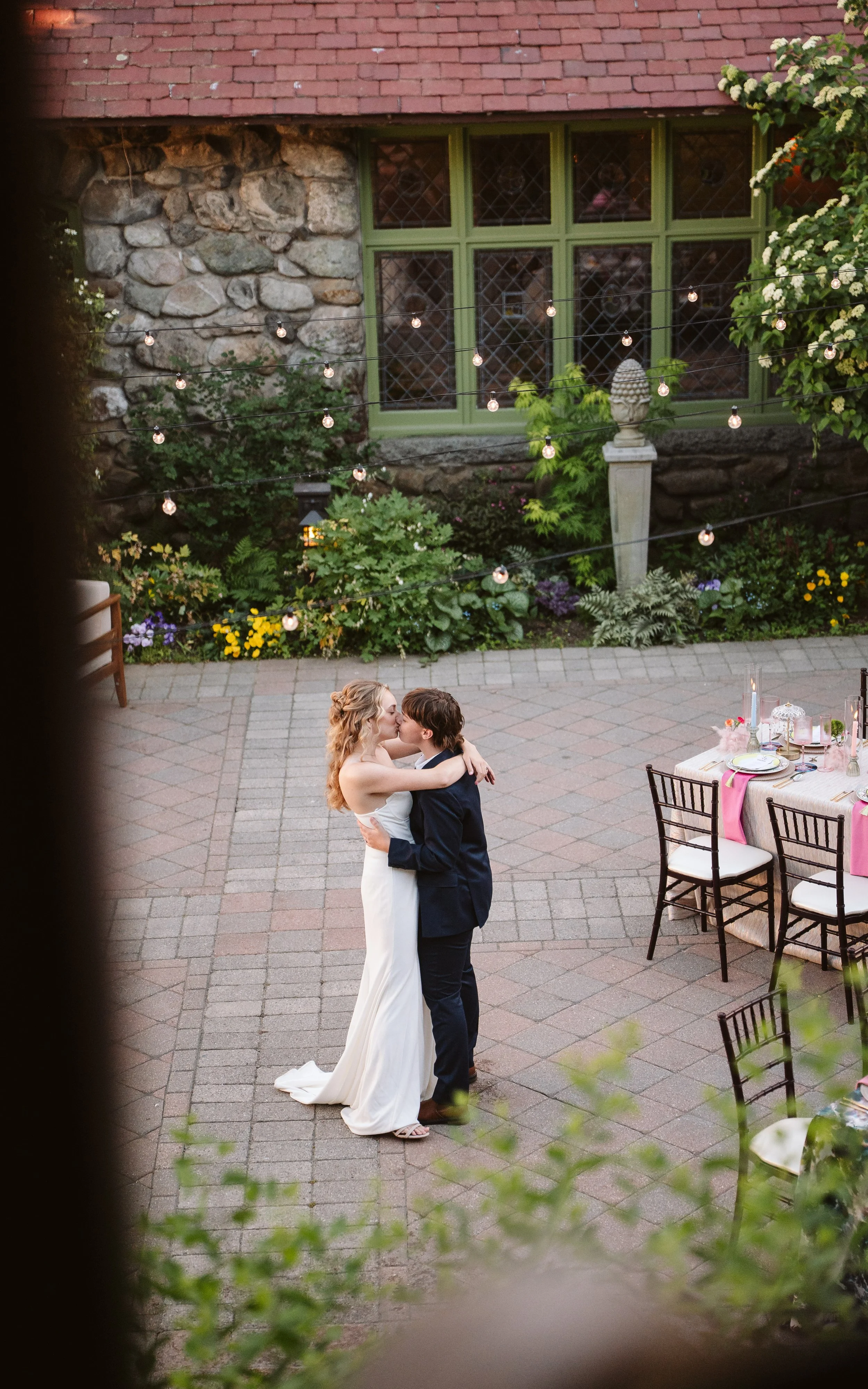 A bride and groom sharing a kiss and embracing outdoors during a wedding reception, with decorated tables and string lights overhead.