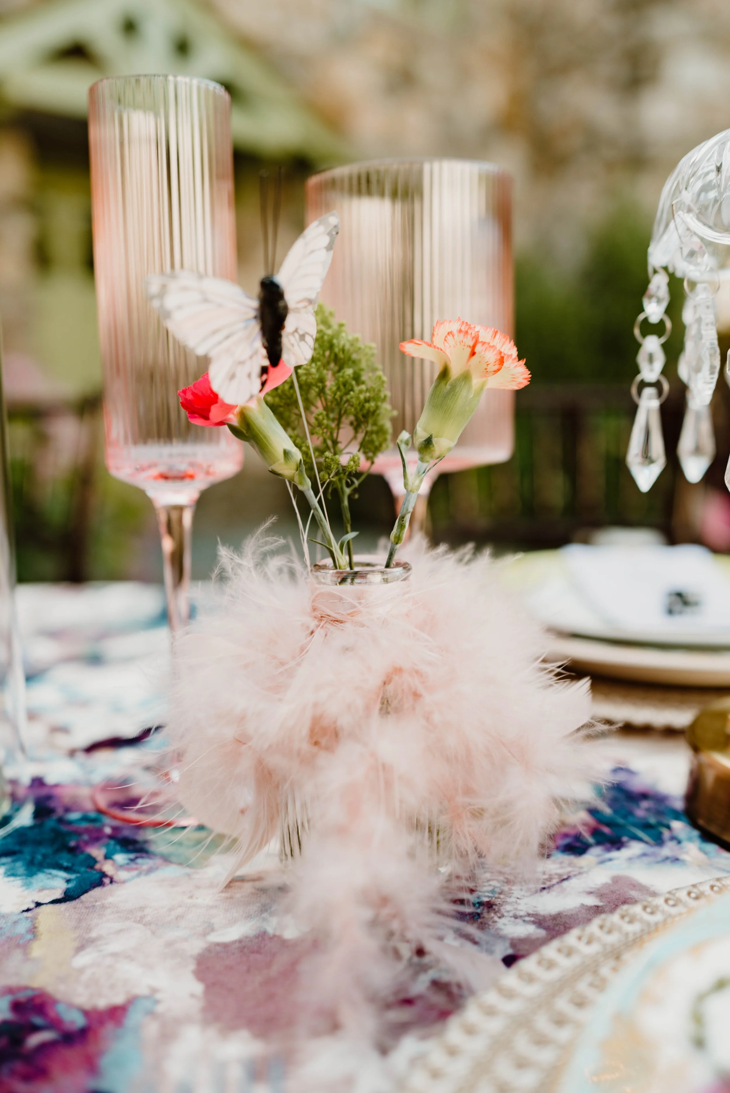 A small glass vase with pink and orange flowers and a white butterfly with black markings, decorated with pink feathers, on a colorful tablecloth.