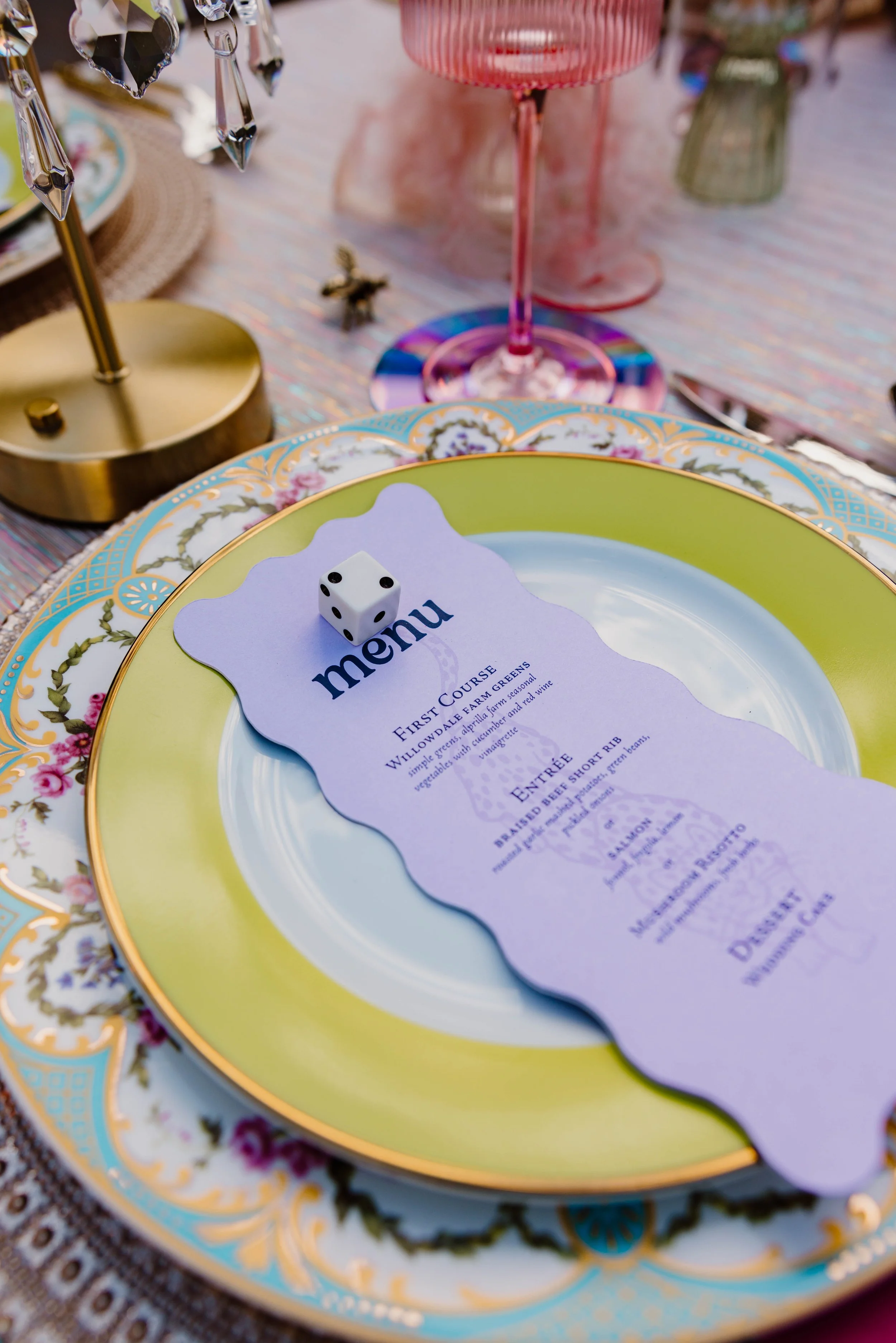 Fancy table setting with a decorative plate, a menu, a pink beverage in a glass, and a gold stand with hanging crystal decorations.