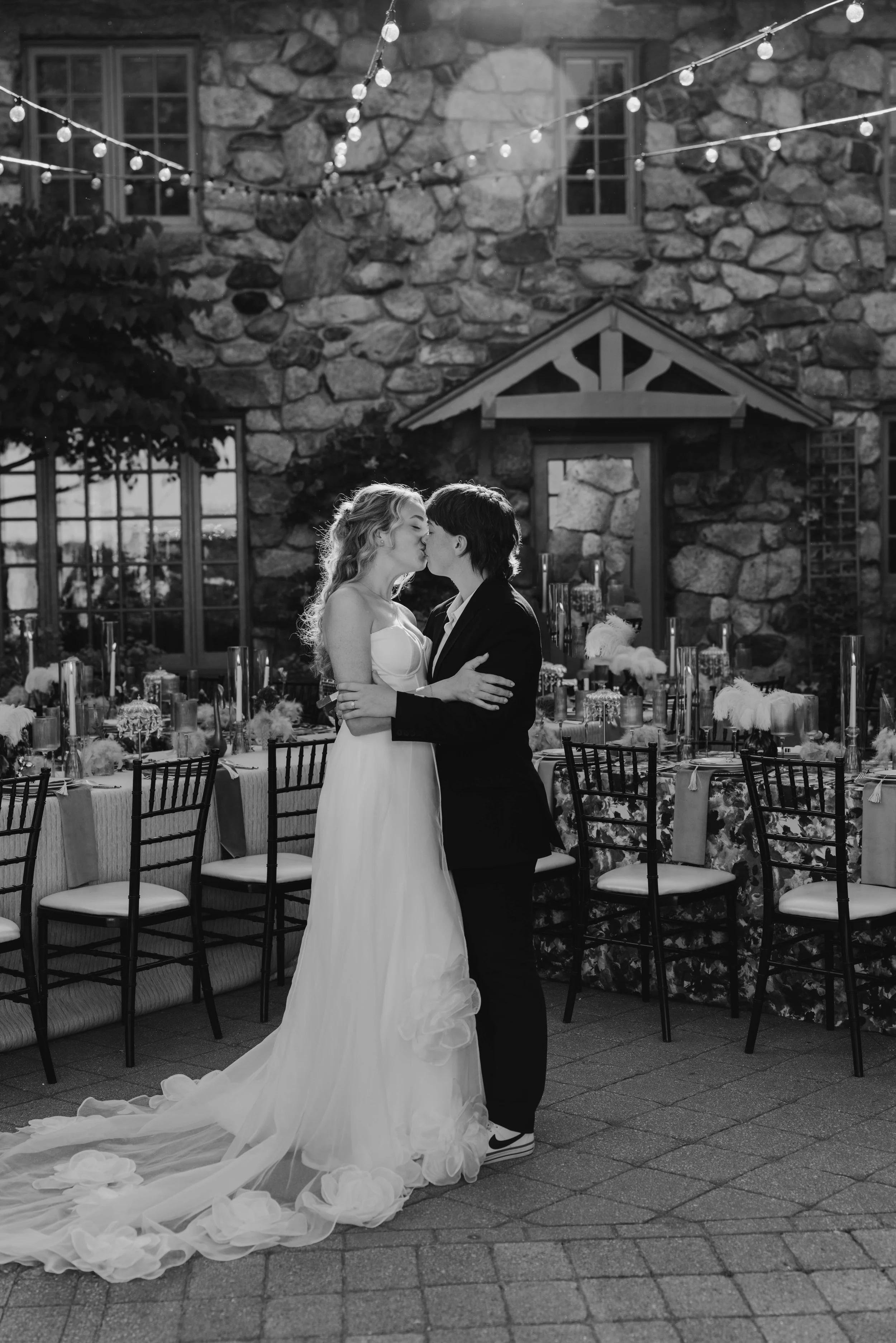 A couple in wedding attire sharing a kiss at an outdoor wedding reception with decorated tables and string lights, against a stone building background.