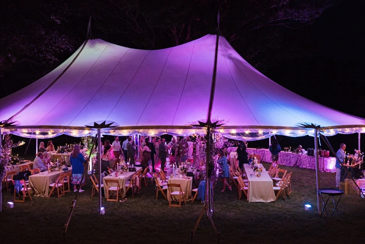 Outdoor nighttime event under a large illuminated tent with people dancing and sitting at tables decorated with flowers.