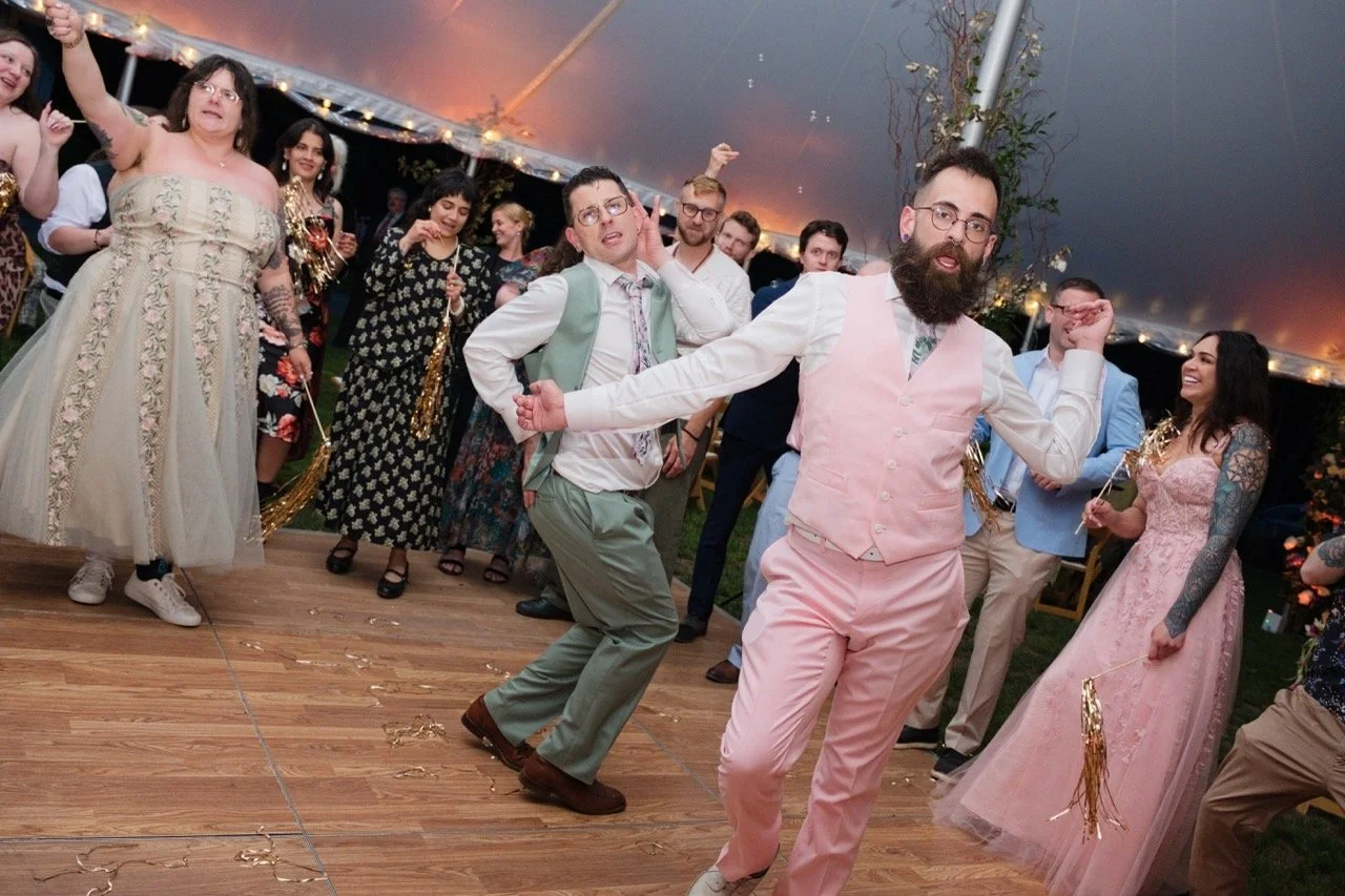People dancing at a wedding reception under a tent with string lights, some holding gold party decorations.